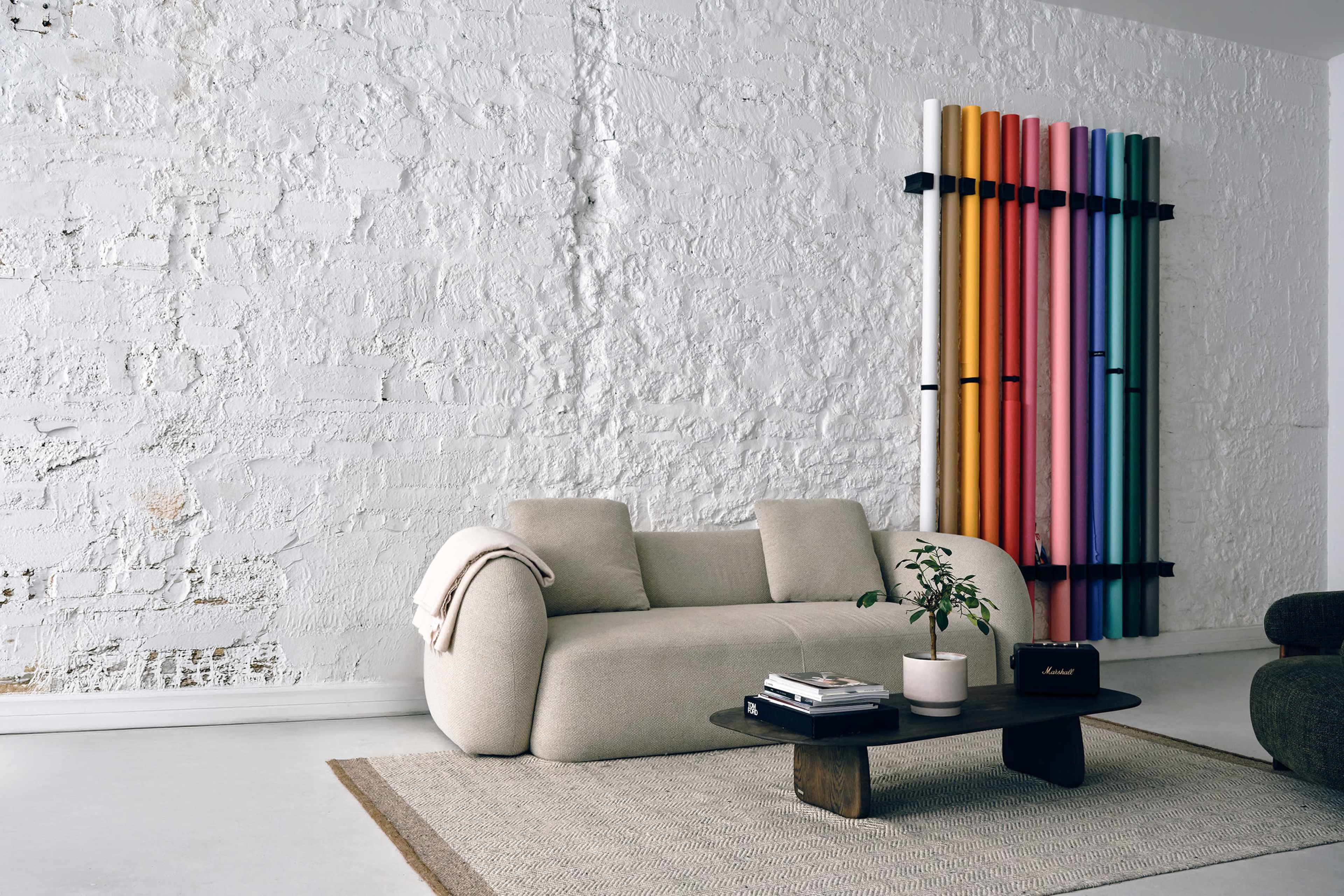 The image shows a modern living room featuring a beige sofa, a round coffee table, a plant, and colorful vertical tubes mounted on a white brick wall.