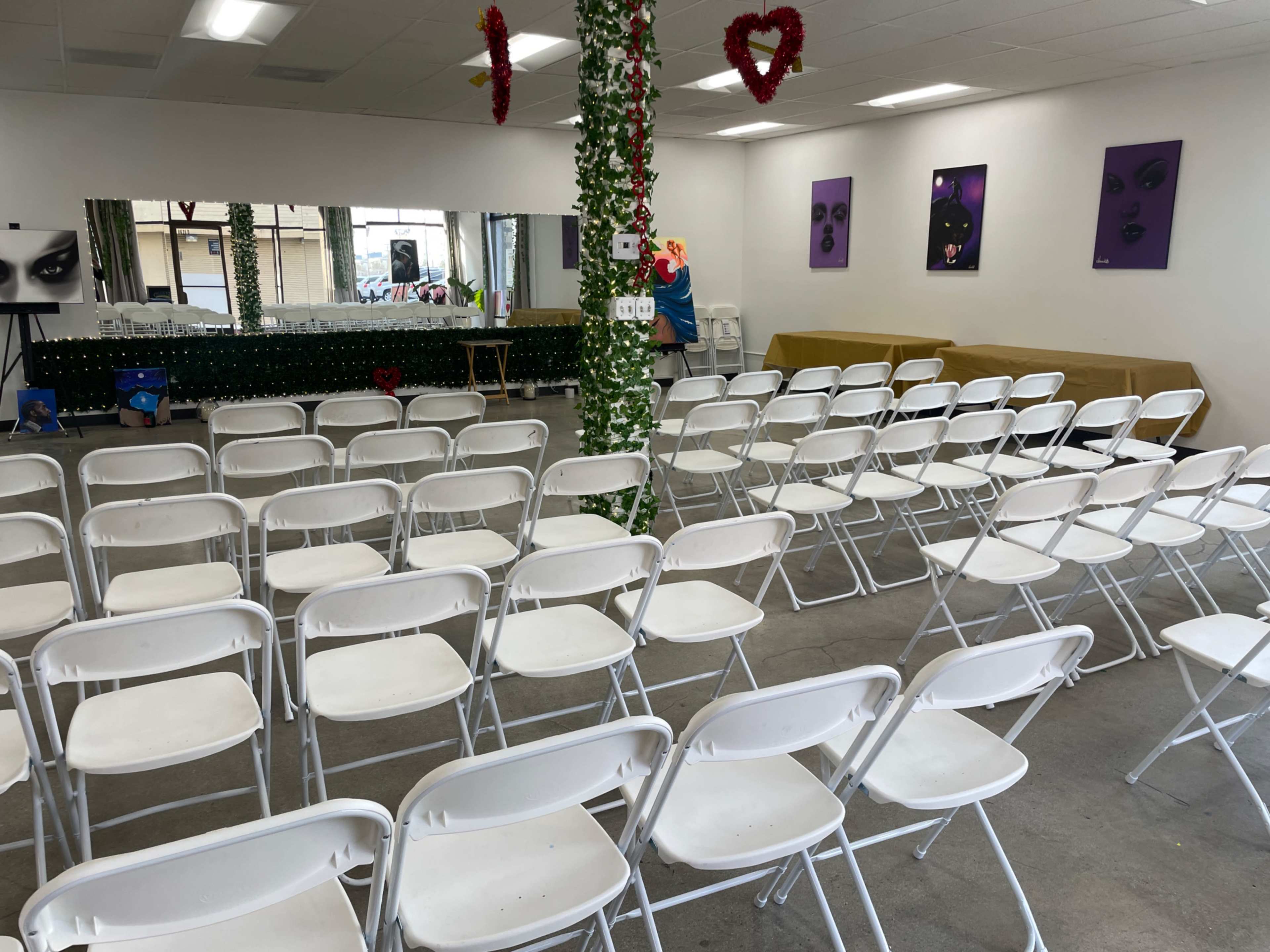 A row of white folding chairs arranged in a clear space, with a mirror and purple artwork on the walls.