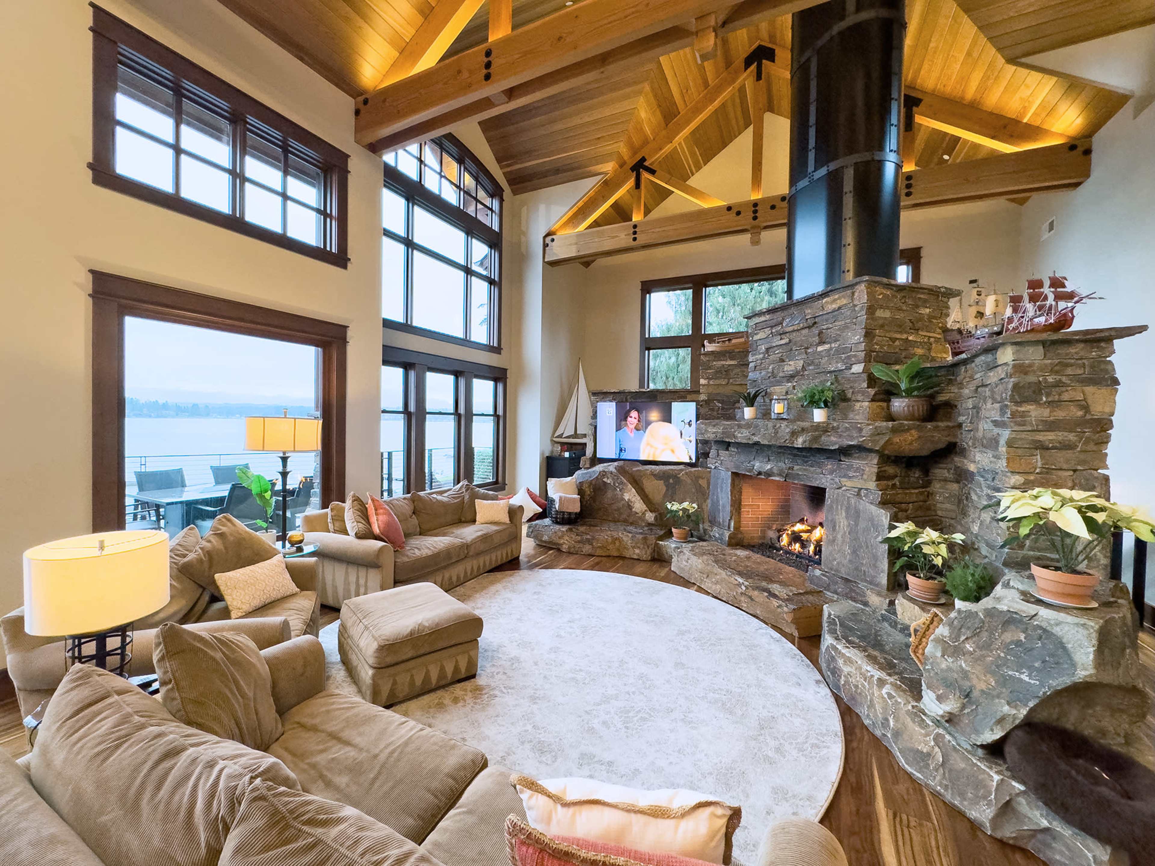 The spacious living room features a circular seating arrangement around a stone fireplace, with large windows providing a view of the lake outside.