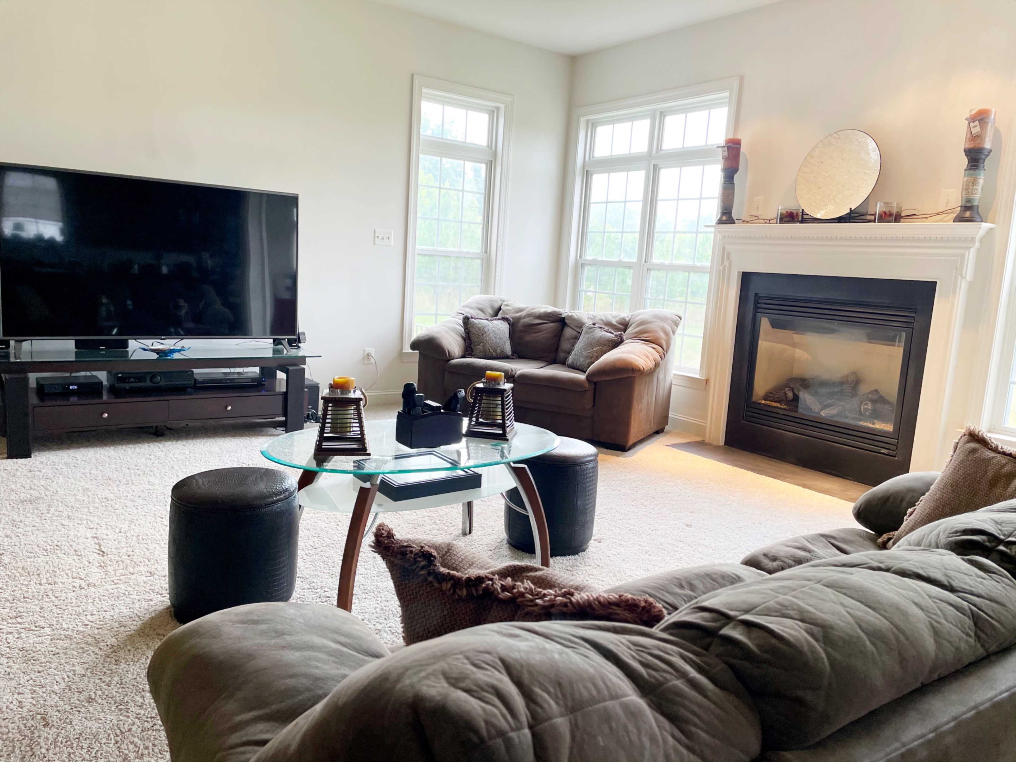 A living room features a large television on a stand, a brown couch, a glass coffee table with decorative items, and a fireplace beside large windows.