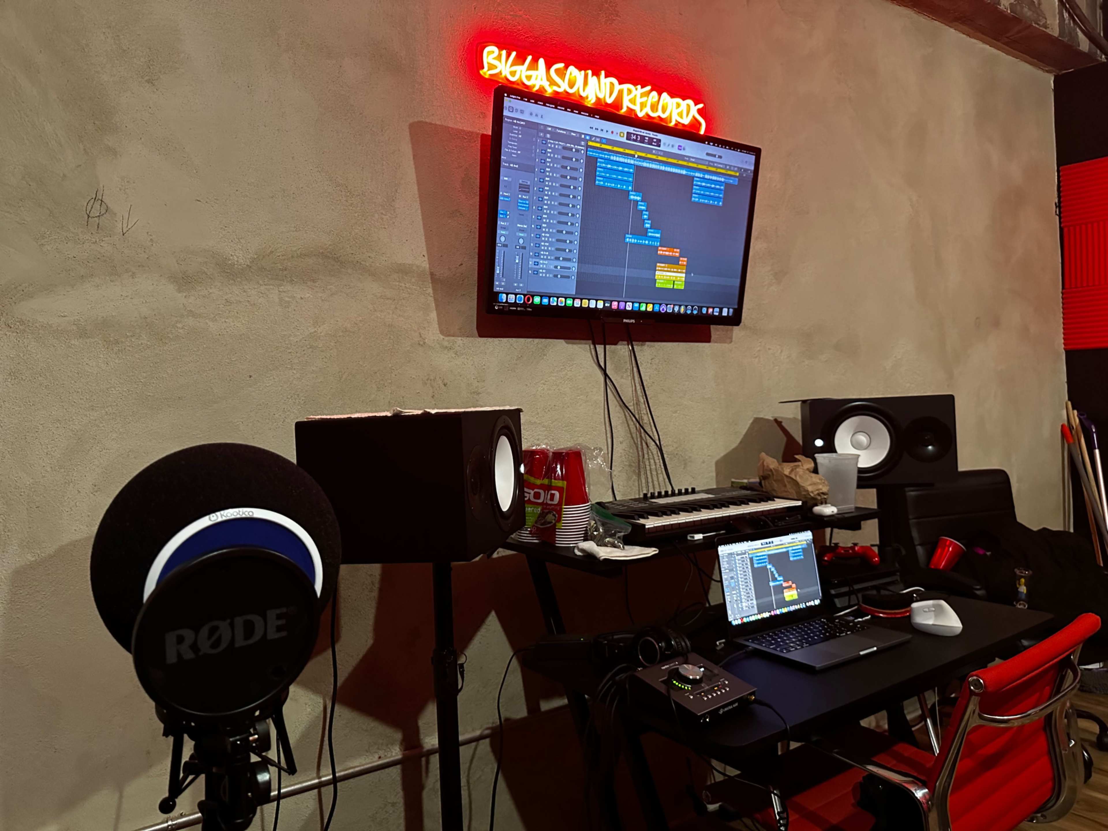 A recording studio setup features a microphone, a computer, and speakers, with a monitor displaying audio editing software and a neon sign overhead.