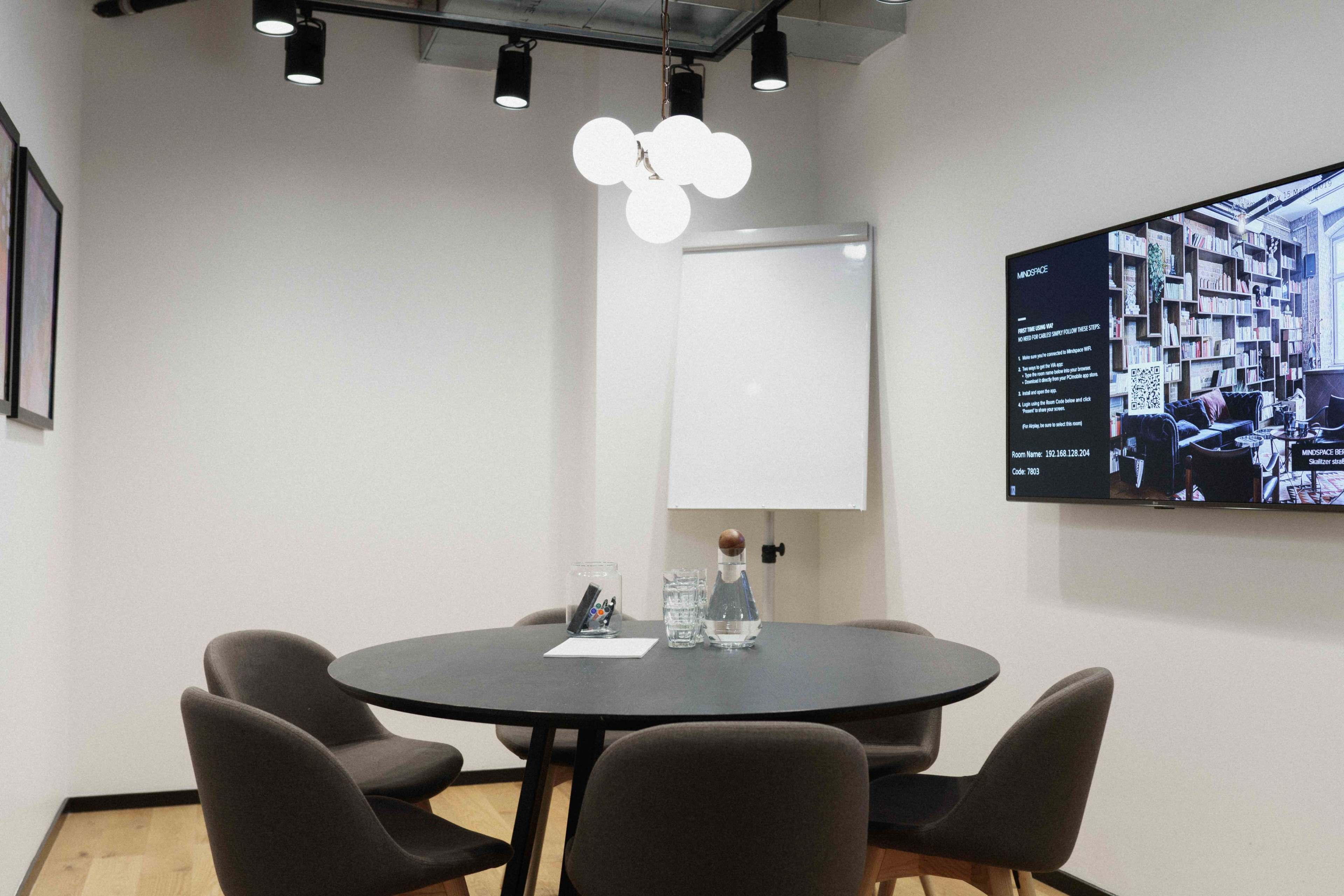 A small meeting room features a round table with four chairs, a whiteboard, and a wall-mounted screen displaying information.