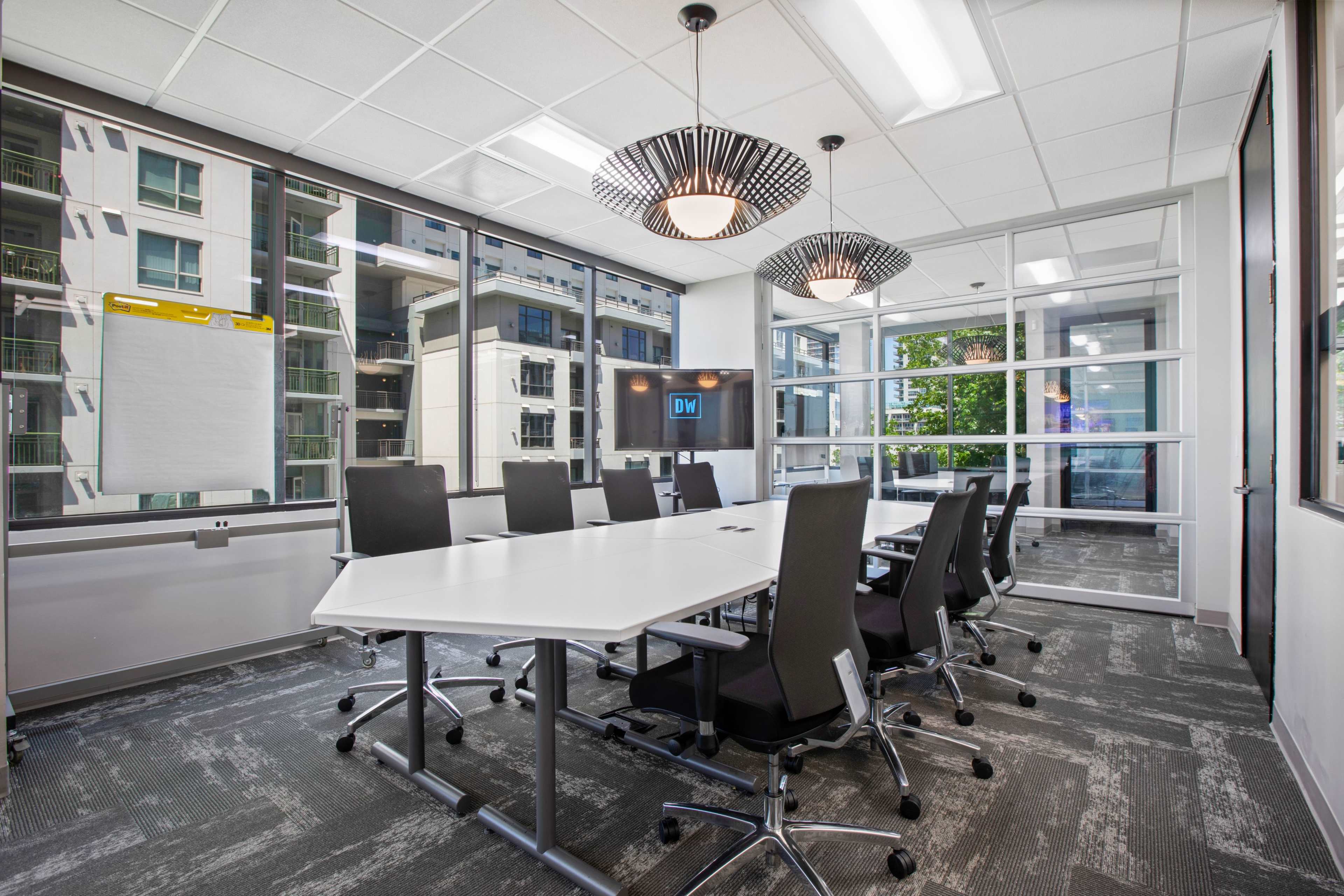 A modern conference room features a large white table surrounded by black chair, with windows providing natural light and a view of the outside.