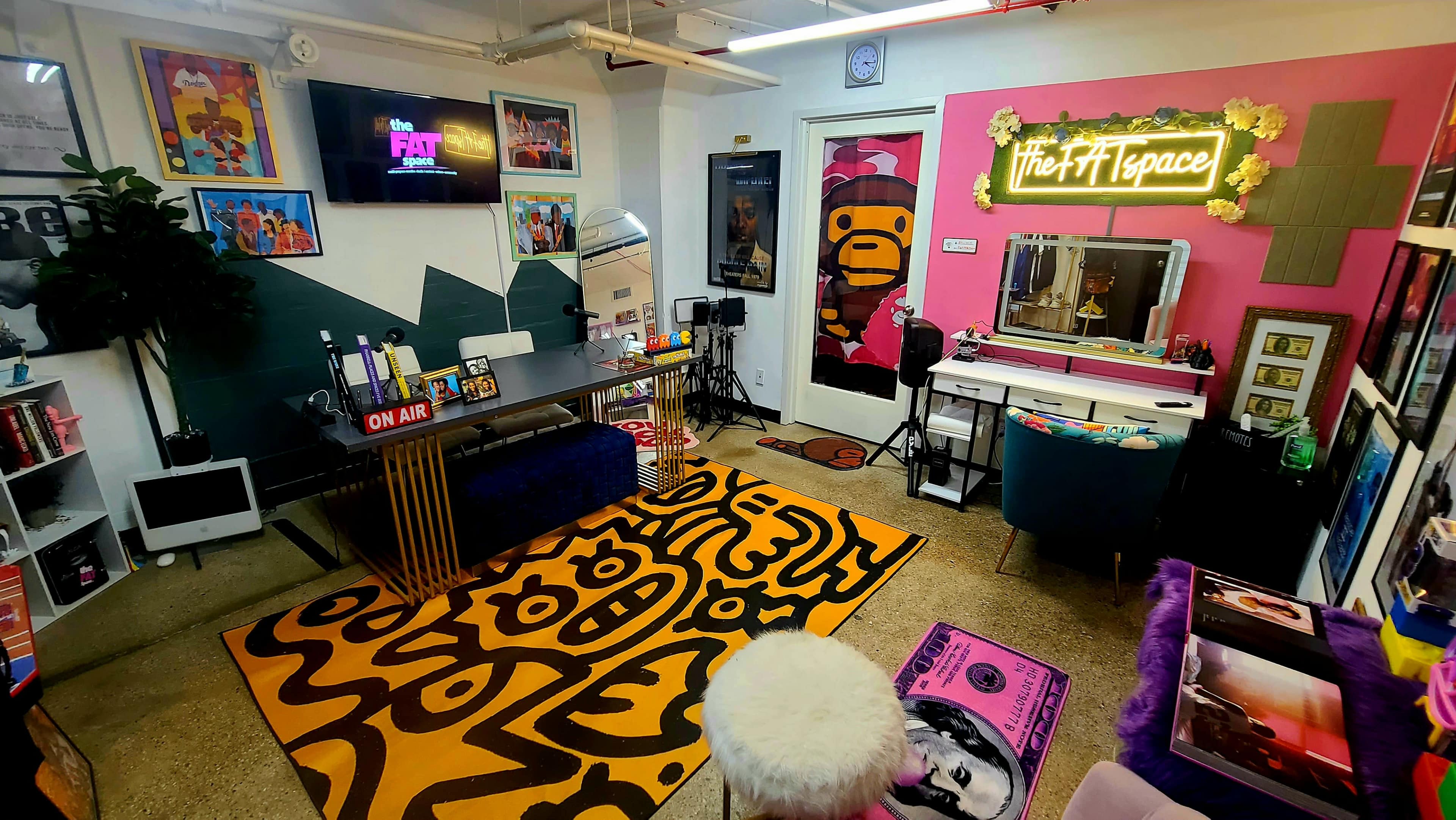 The image shows a colorful indoor creative space with a large yellow patterned rug, a vanity area, and a wall featuring a bright pink sign that reads "The FTF Space."