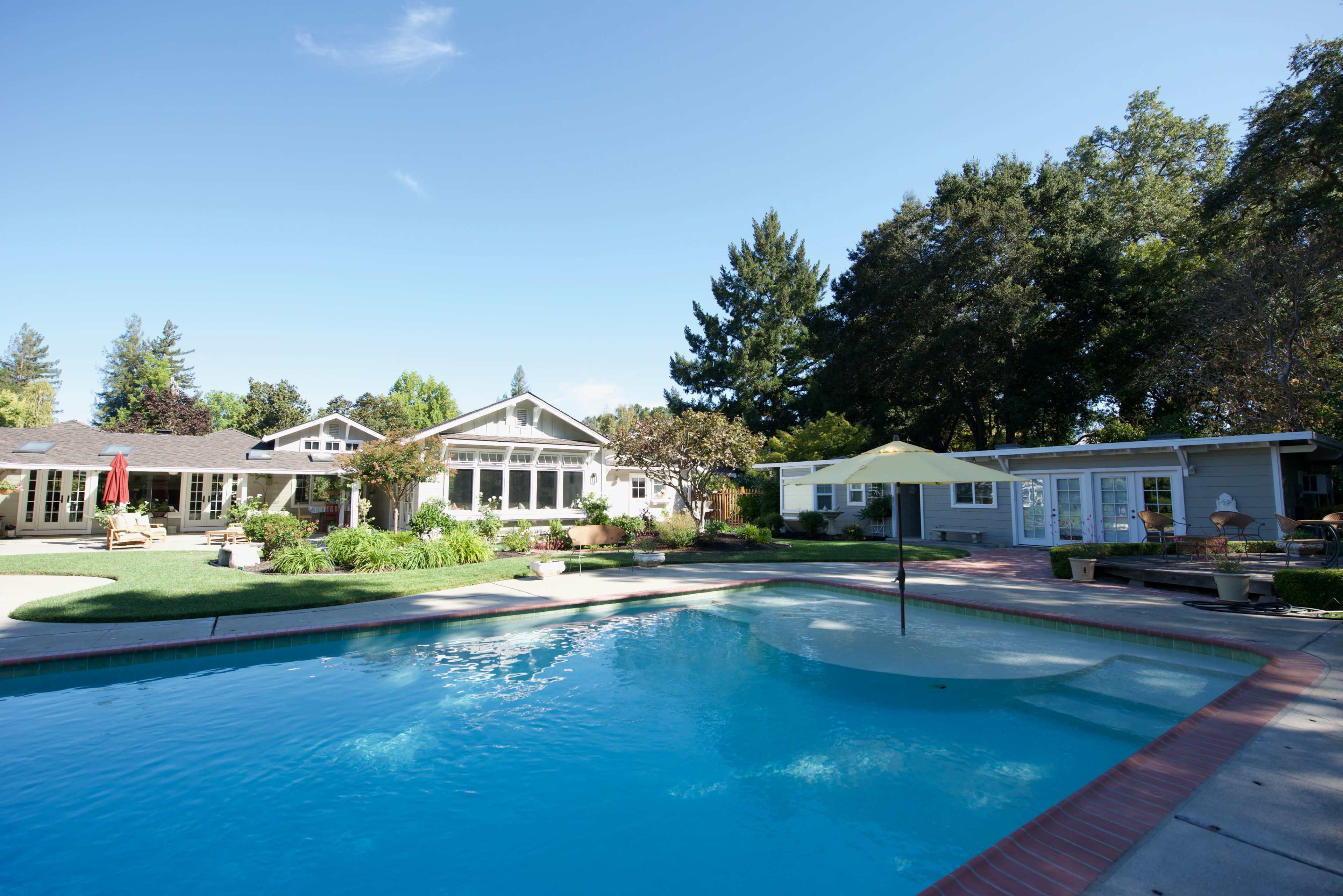 A residential backyard features a swimming pool surrounded by green grass, trees, and multiple houses.