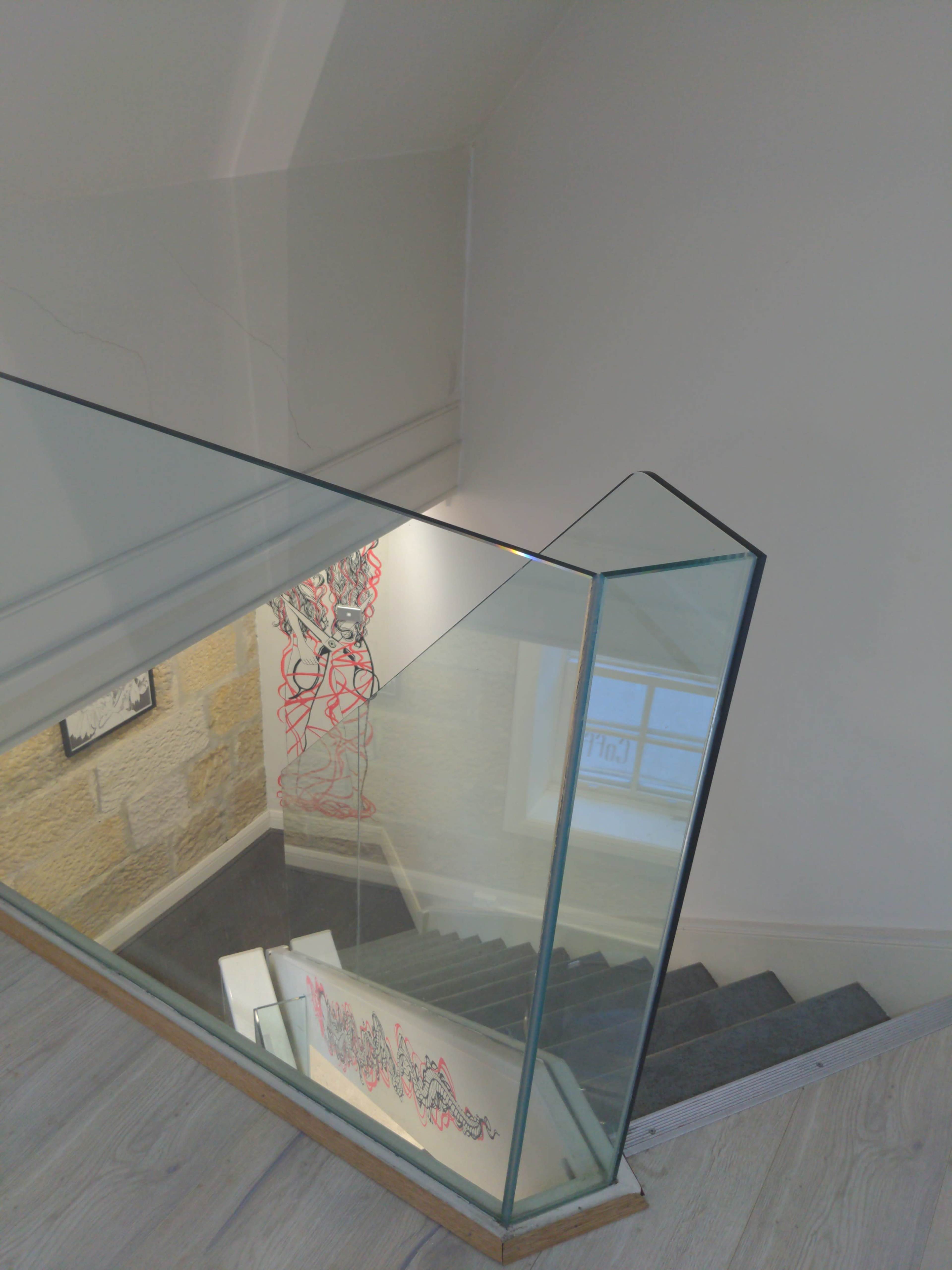 The image shows a glass railing overlooking a stairway with light-colored wooden steps, leading down to a room with artwork visible on the walls.