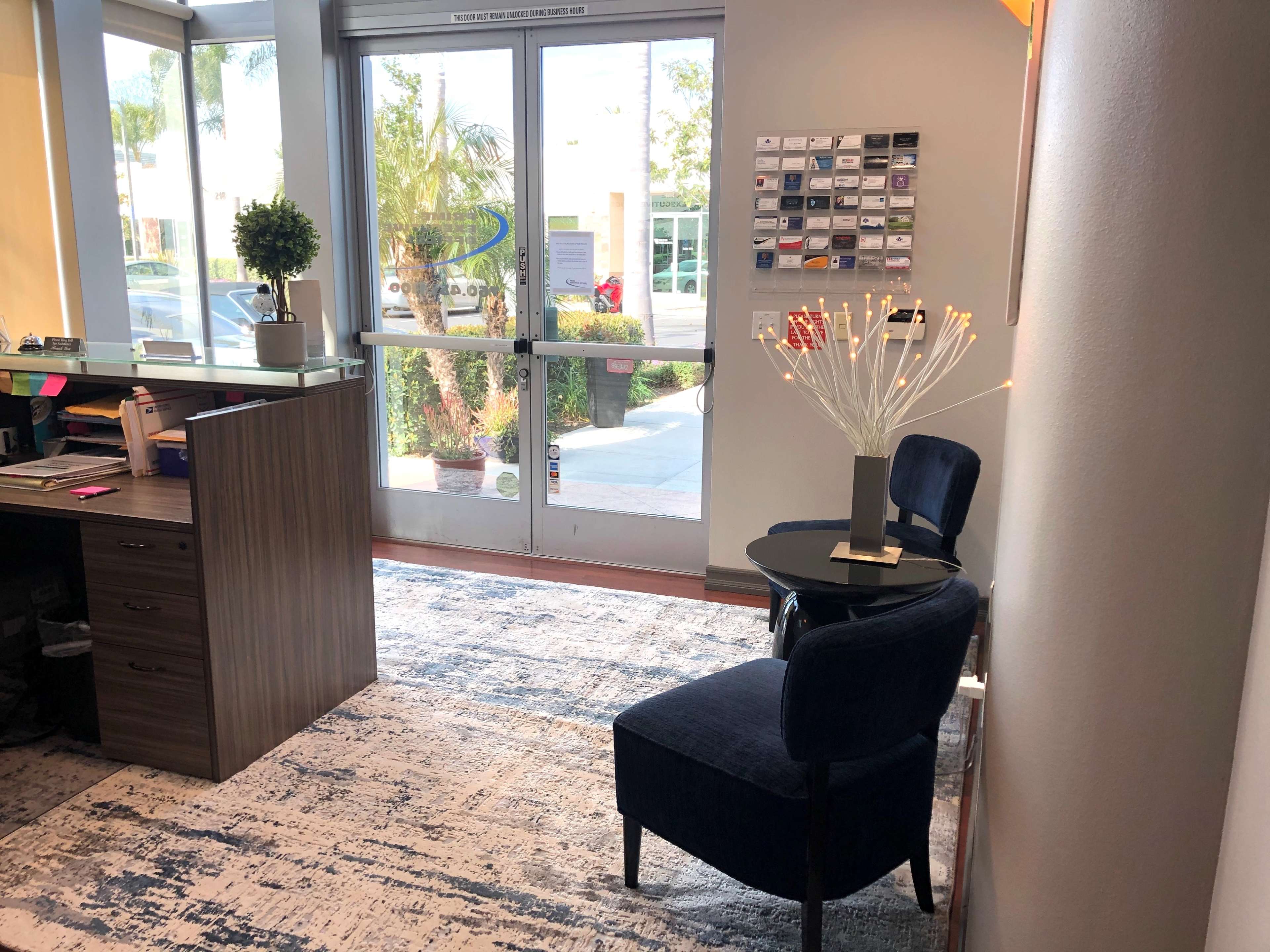 The image shows an office space with a reception area featuring a desk, two blue chairs, a decorative plant, and a wall display of various business cards.