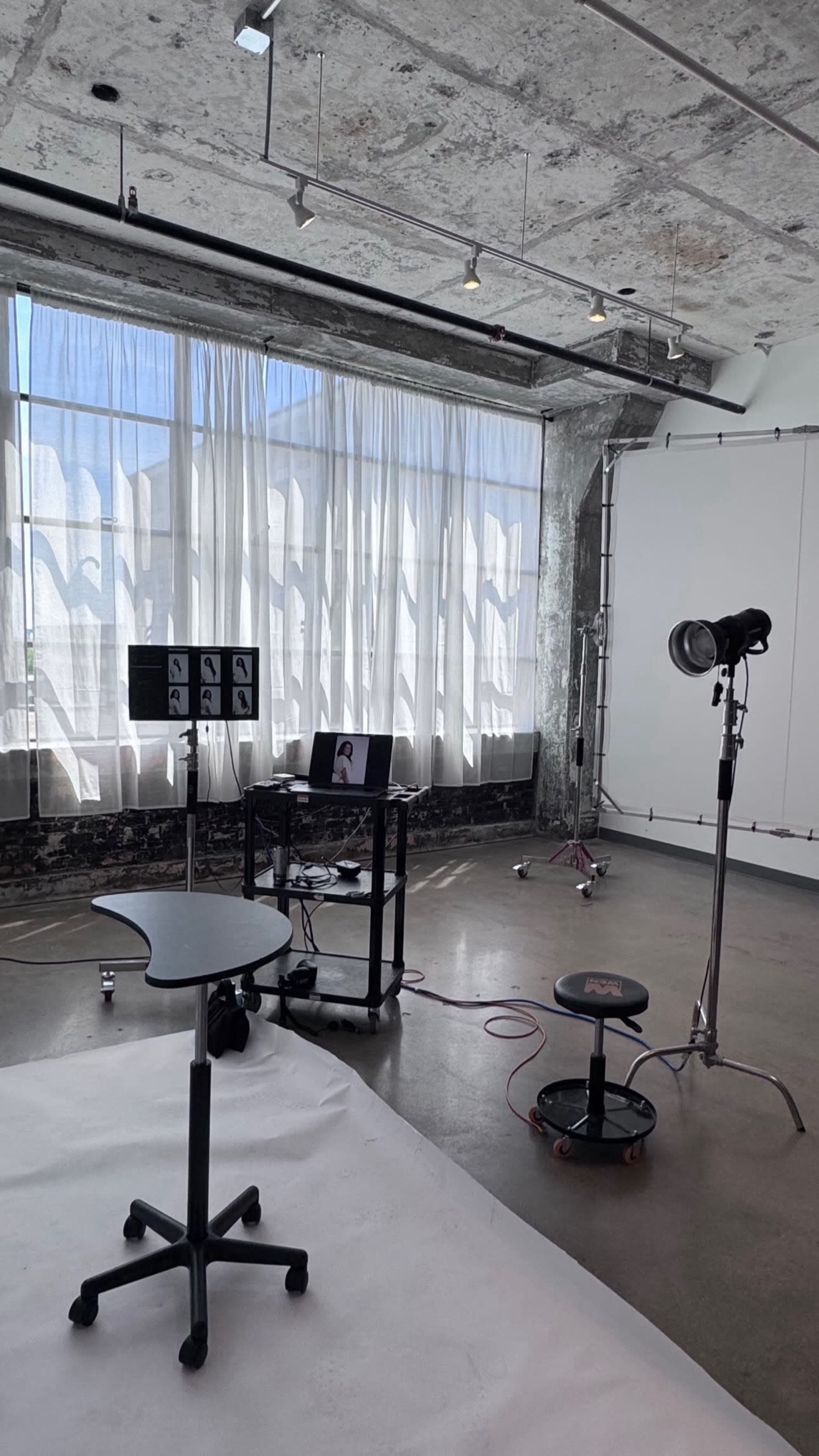 The image shows a spacious photography studio with large windows, a white backdrop, a table with a computer and monitor, and various lighting equipment.
