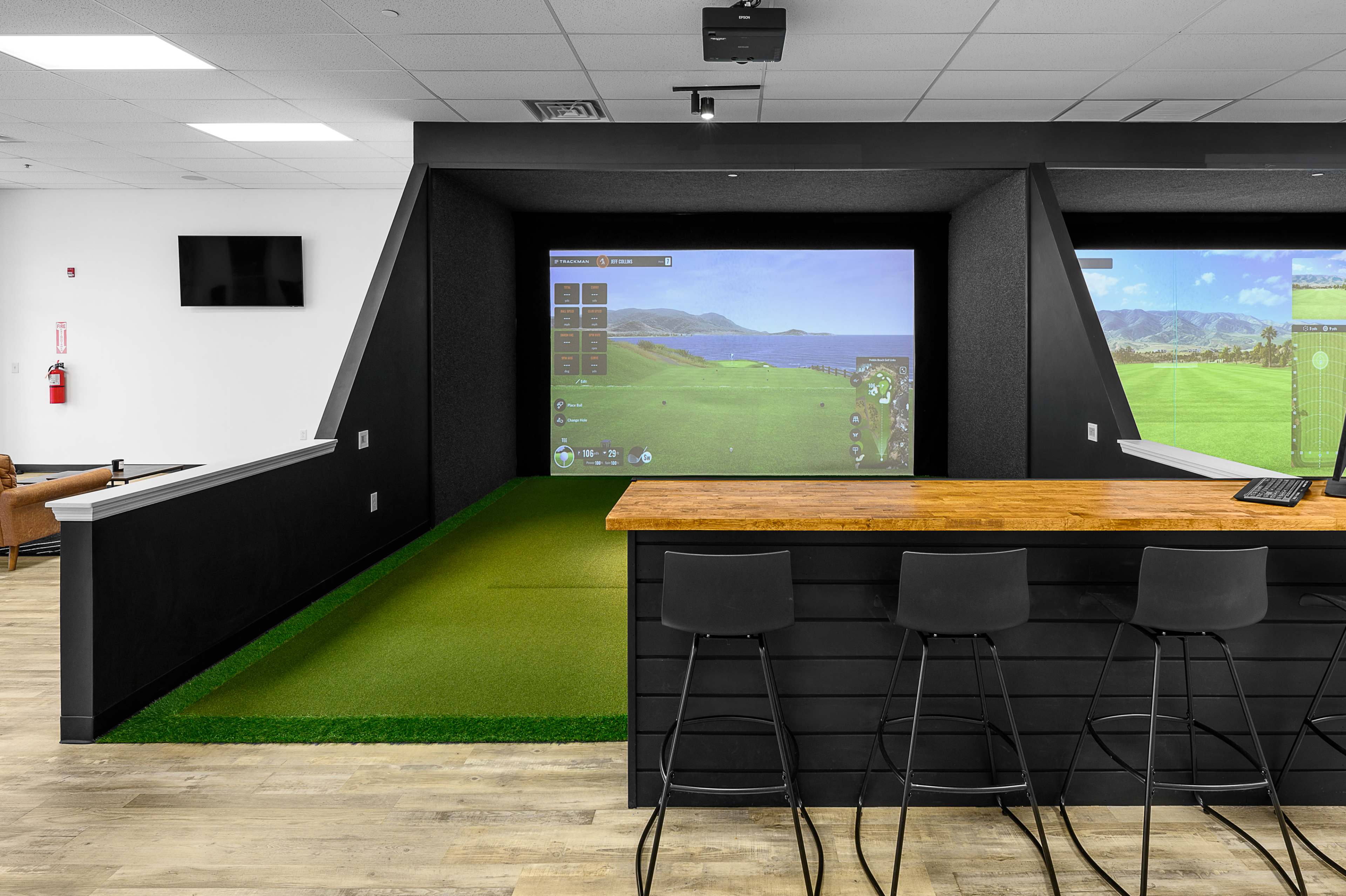 The image shows an indoor golf simulation area with a large screen displaying a virtual golf course and a bar with high stools in the foreground.