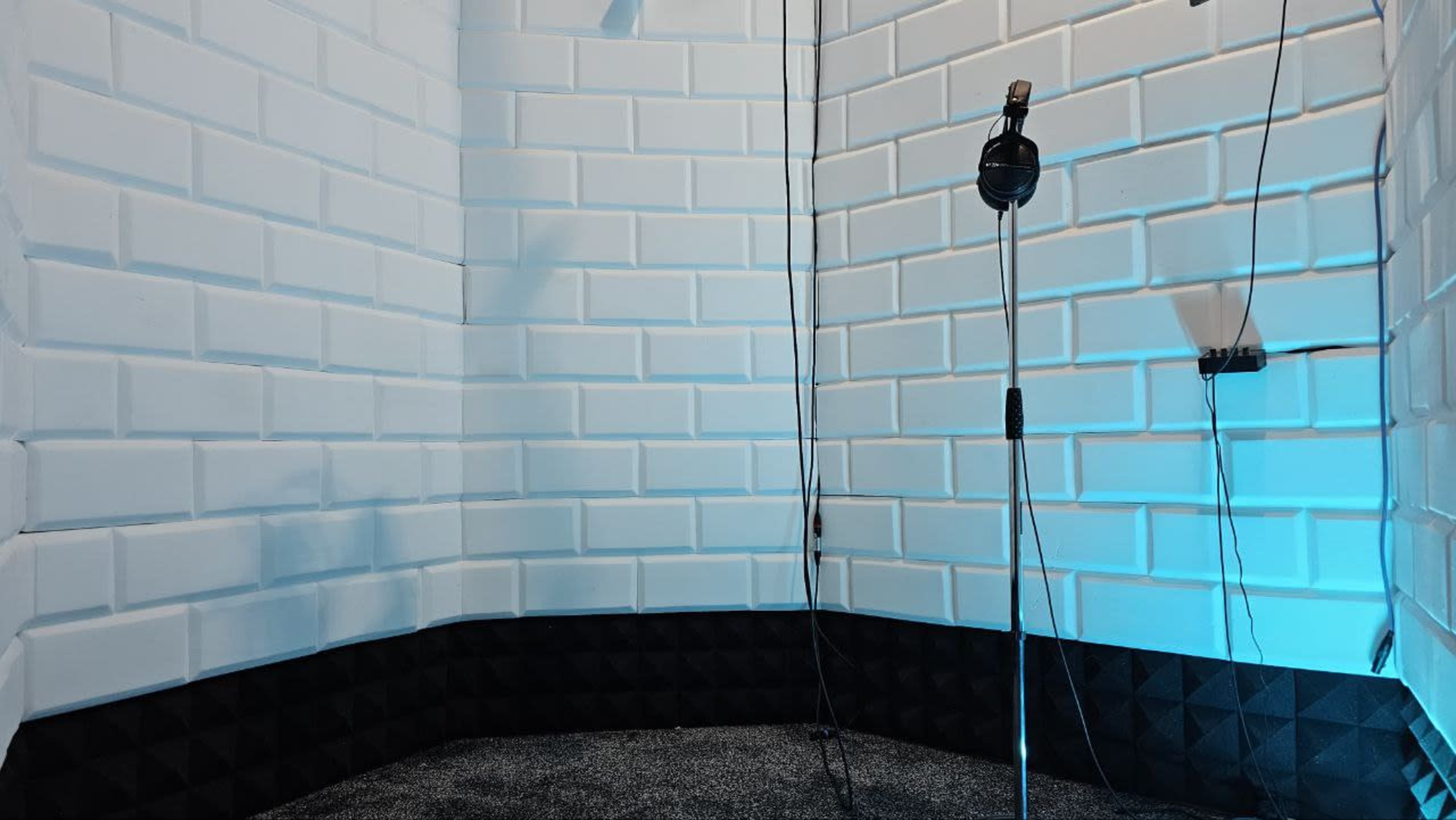 A small soundproof recording booth with white brick walls, a microphone on a stand, and a dark carpeted floor.