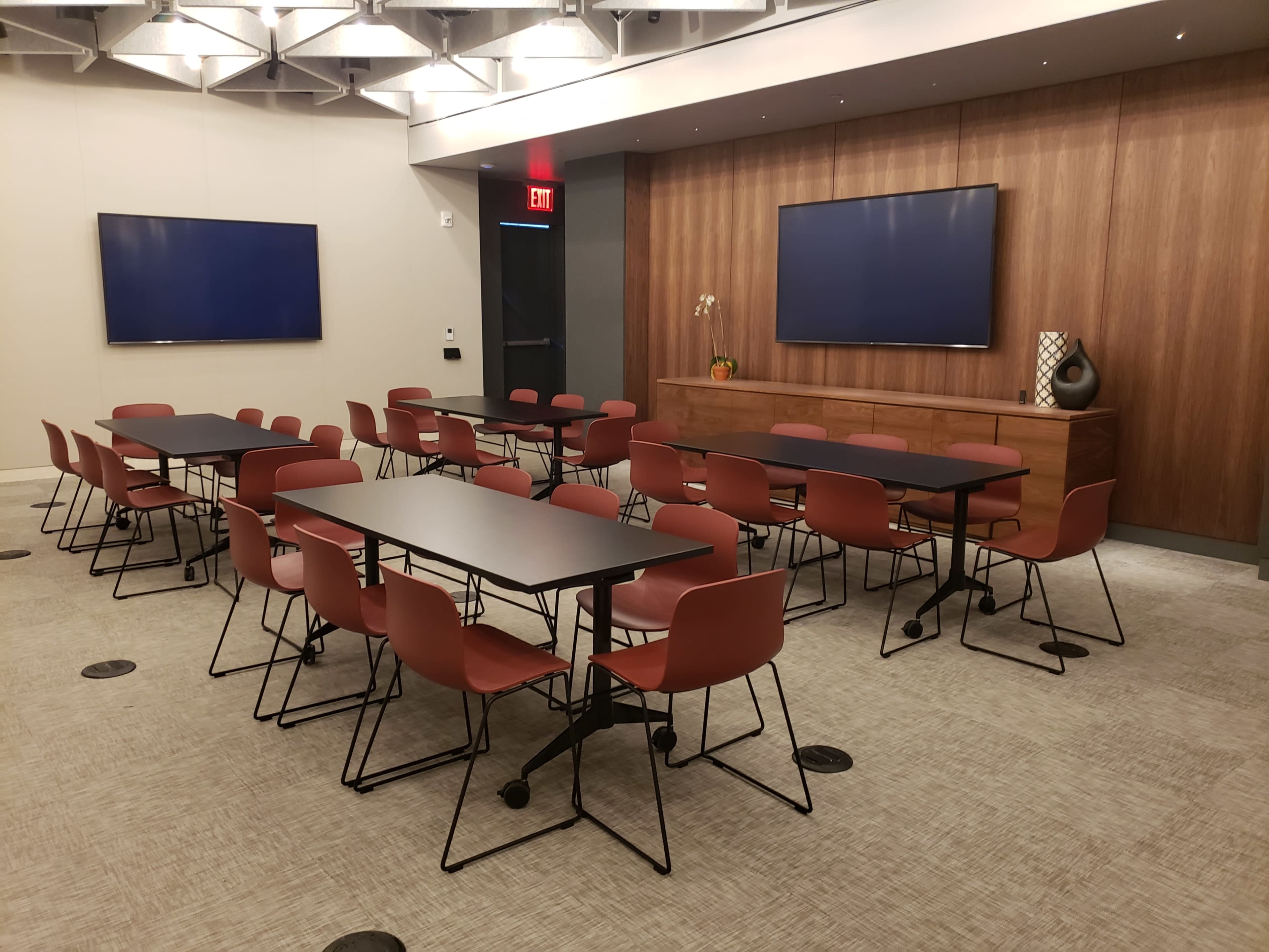 A conference room with multiple black rectangular tables and red stacking chairs arranged in two clusters, featuring two large screens on the walls and a wooden cabinet along the back.