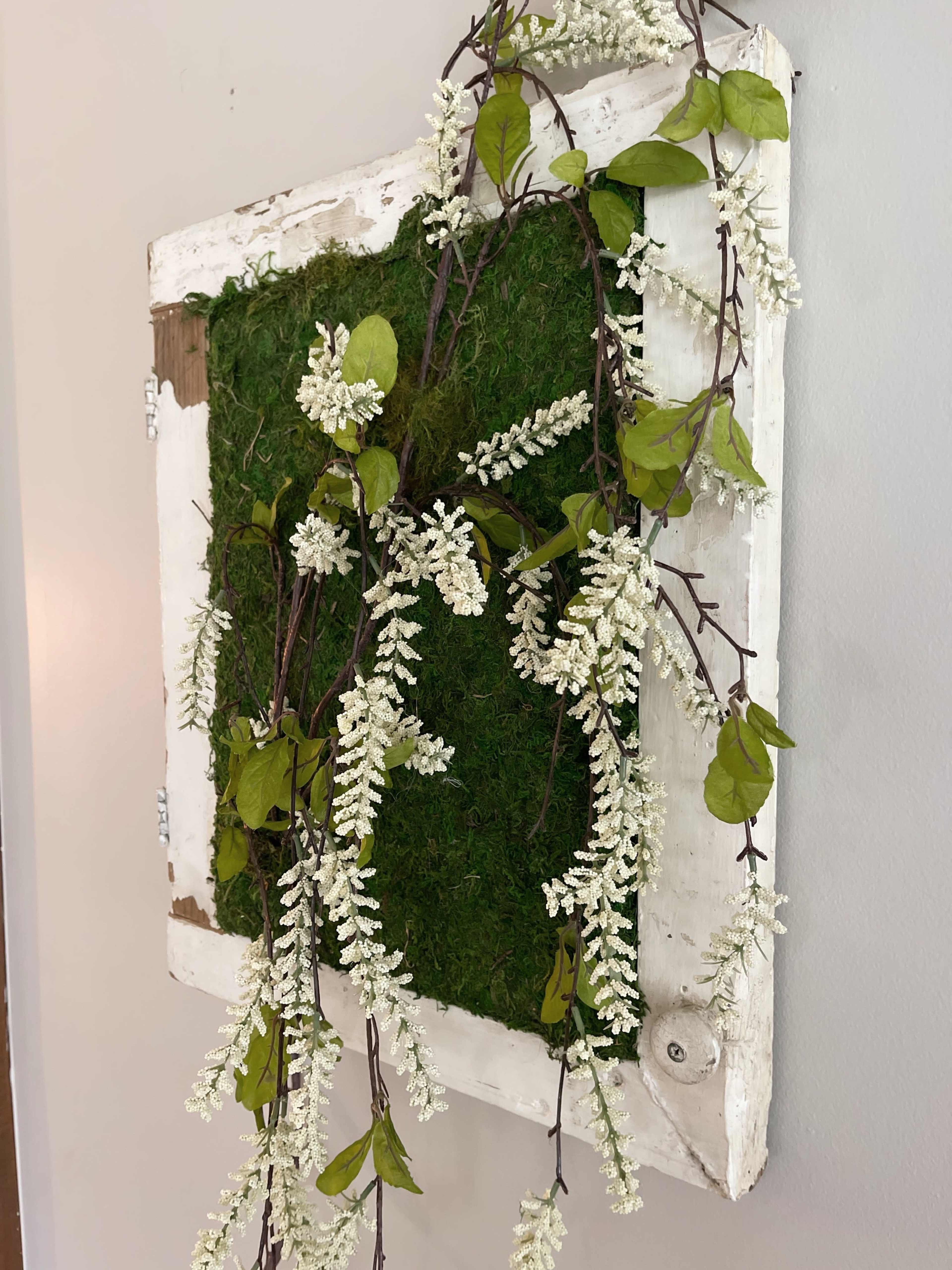 A framed piece of moss art displays white floral accents and green leaves cascading down.