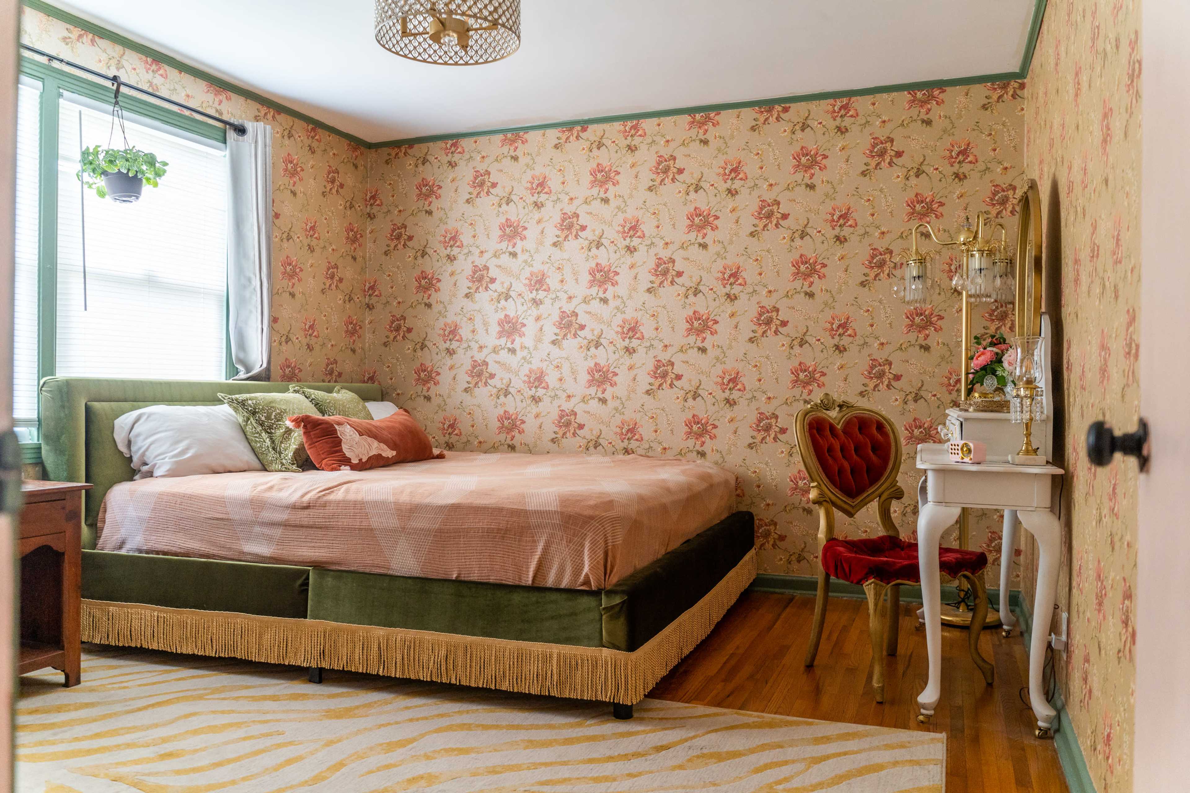 A bedroom with floral wallpaper, a green bed, a wooden nightstand, and a small vanity table with a red chair.