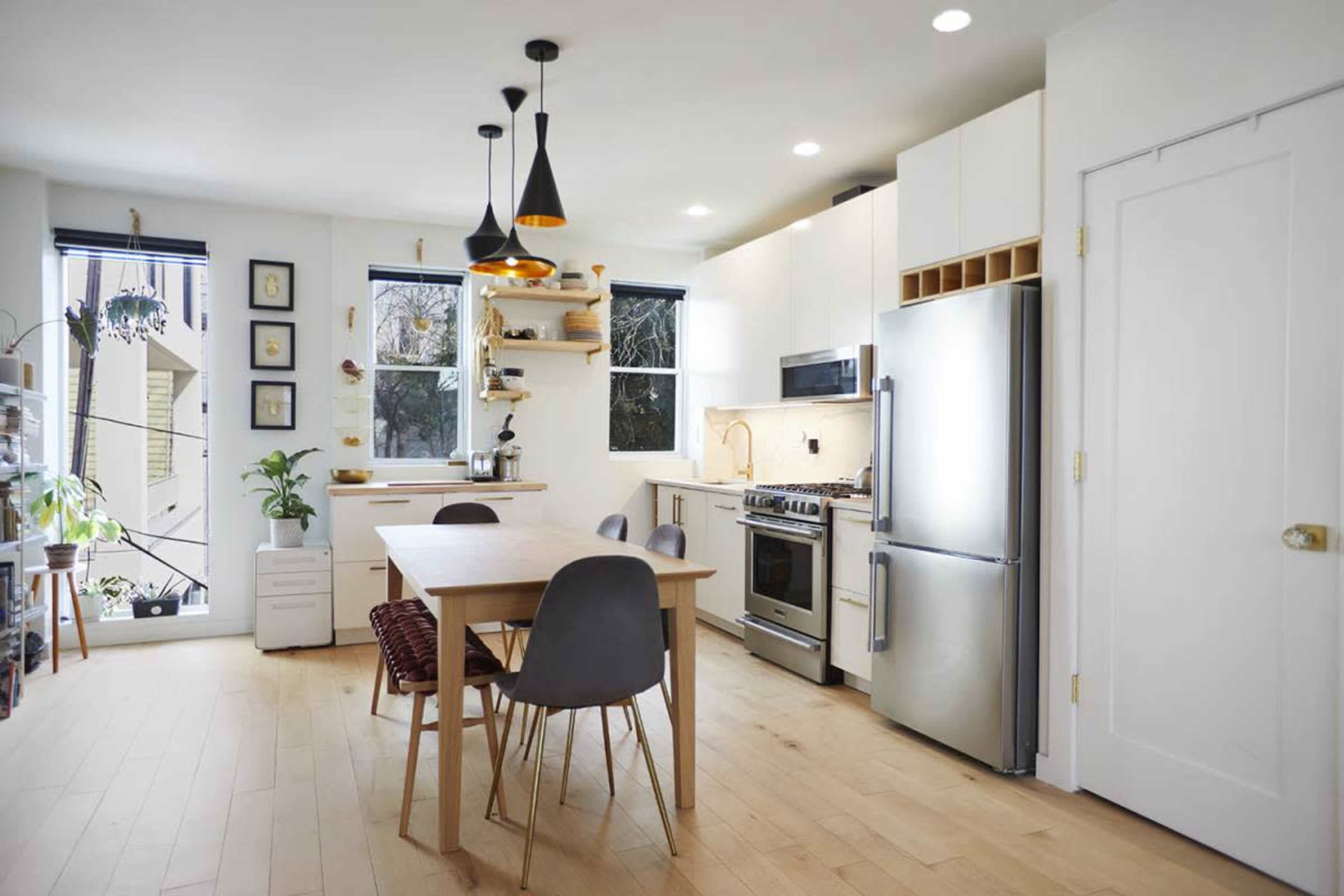 A modern kitchen features a central wooden dining table, sleek white cabinetry, stainless steel appliances, and large windows allowing natural light to flood the space.