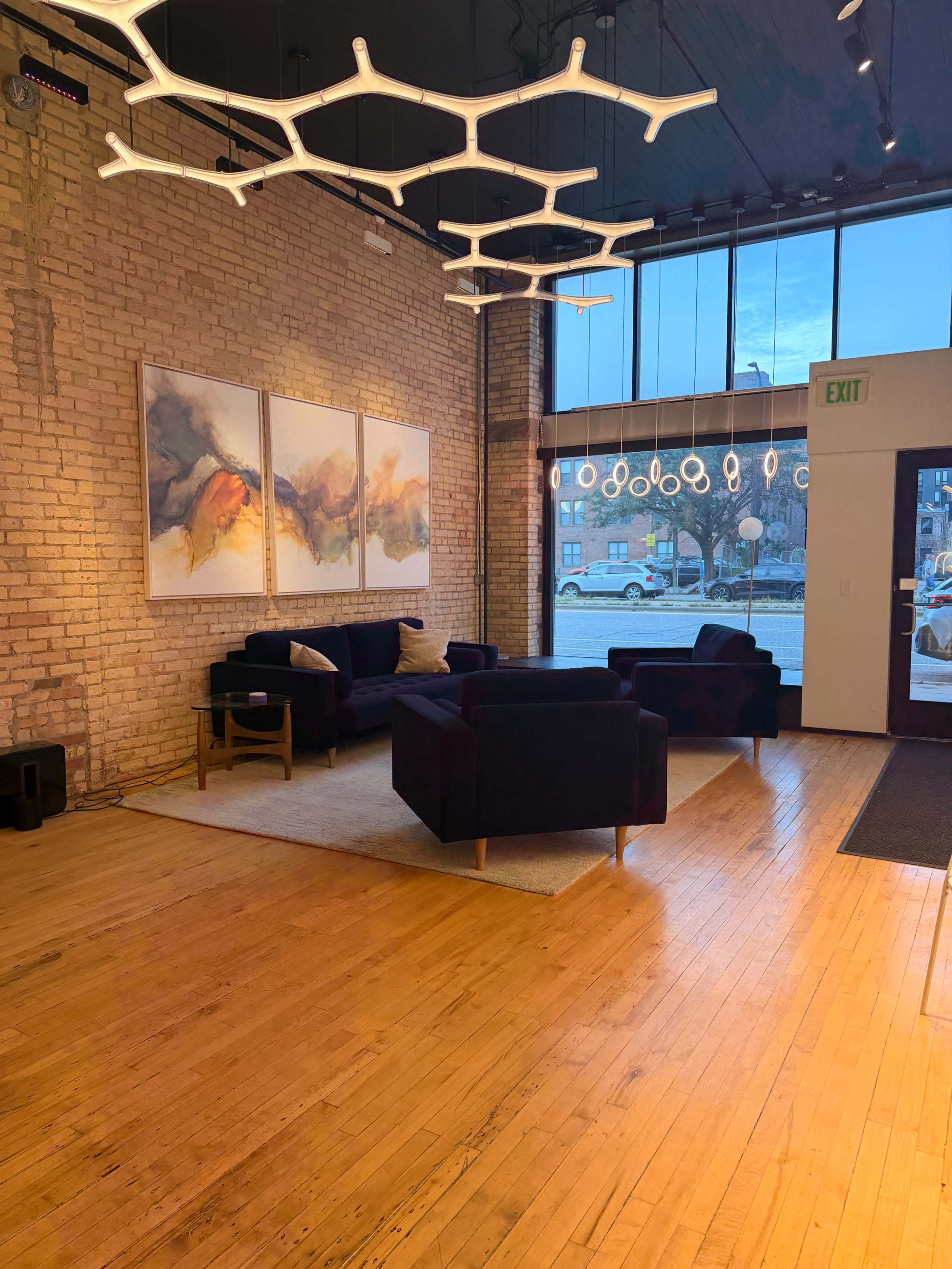The photo depicts a modern lounge area featuring dark armchairs and a large rug, with abstract artwork displayed on the brick wall and large windows letting in natural light.