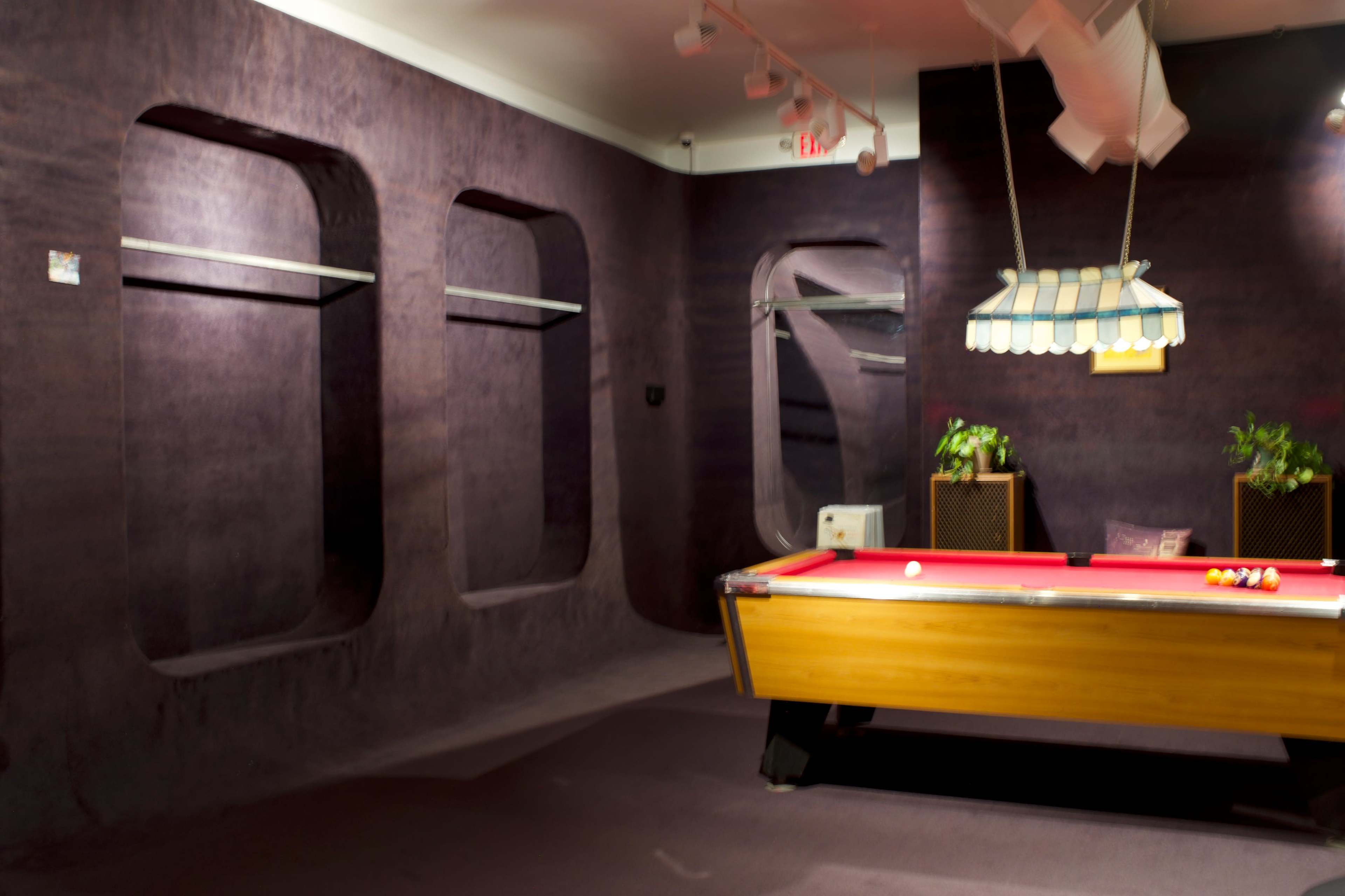 The image shows a billiards room with a red pool table, two small plants in decorative containers, and a few wall niches, all surrounded by purple walls.