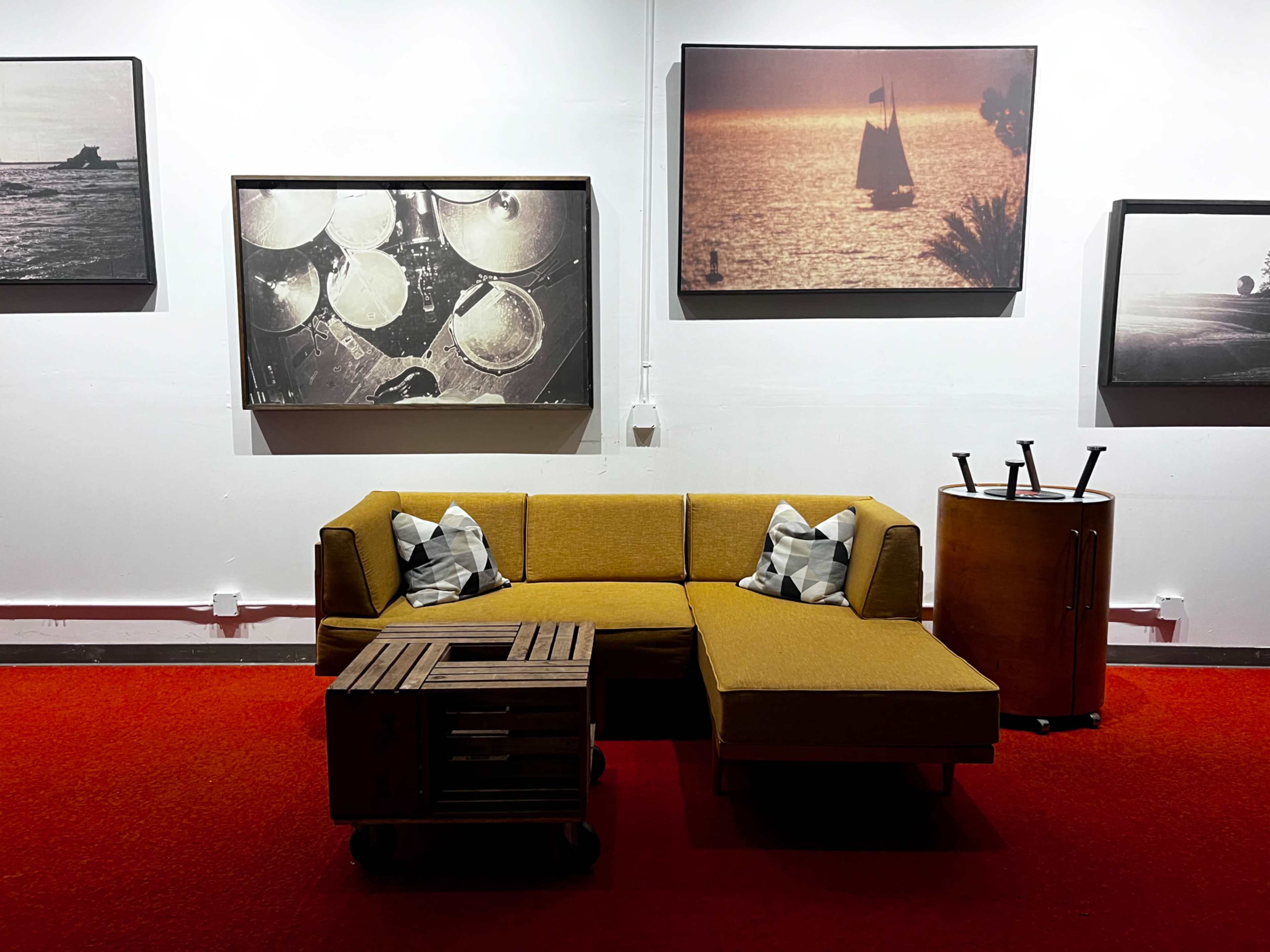 The image shows a yellow sectional sofa with patterned throw pillows, a wooden coffee table, and a round side table, all positioned on a red carpet against a backdrop of framed photographs.