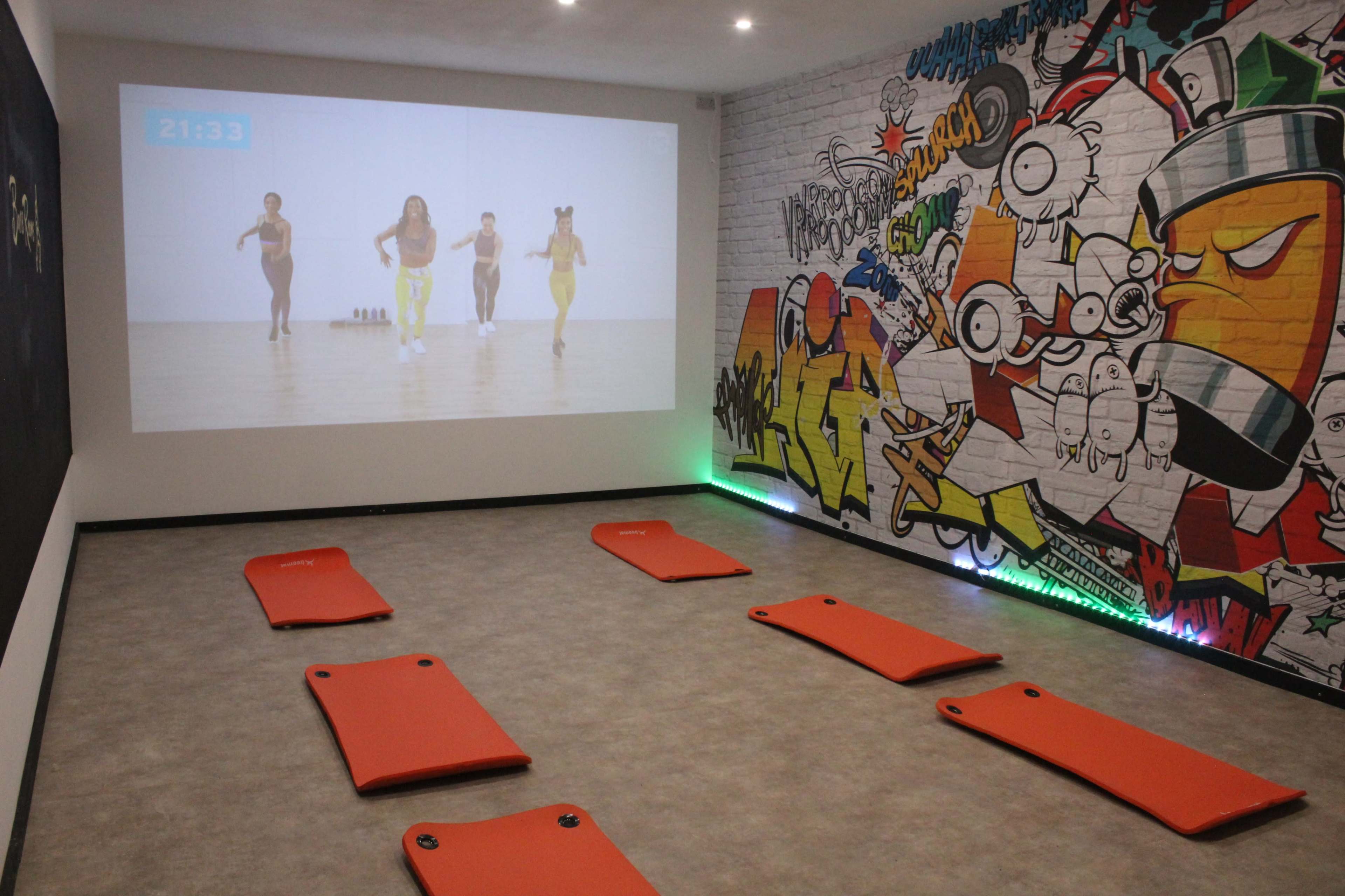 The room features a large screen displaying a workout session, with orange exercise mats arranged on the floor and a vibrant graffiti mural on one wall.