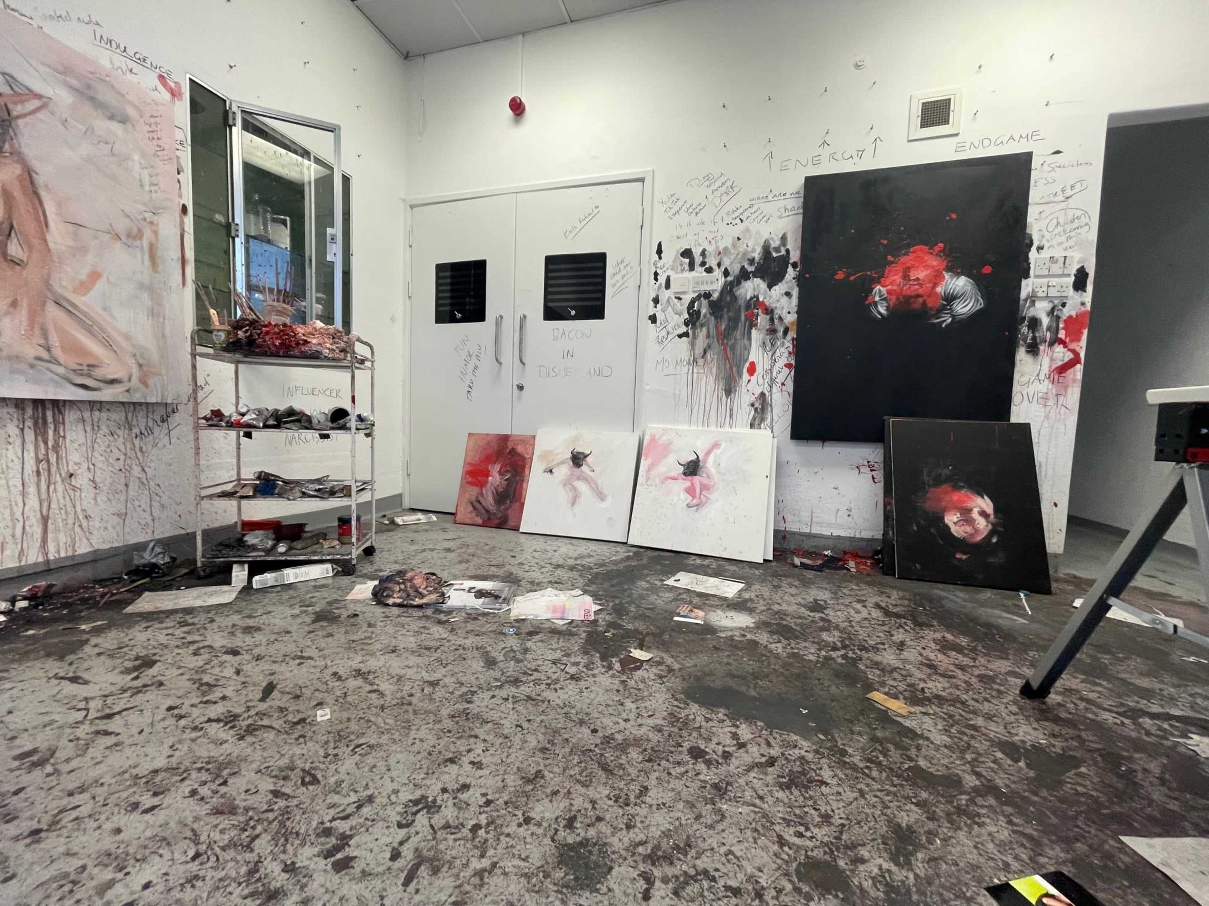 A messy art studio with scattered paintings and a floor covered in paint splatters and debris.
