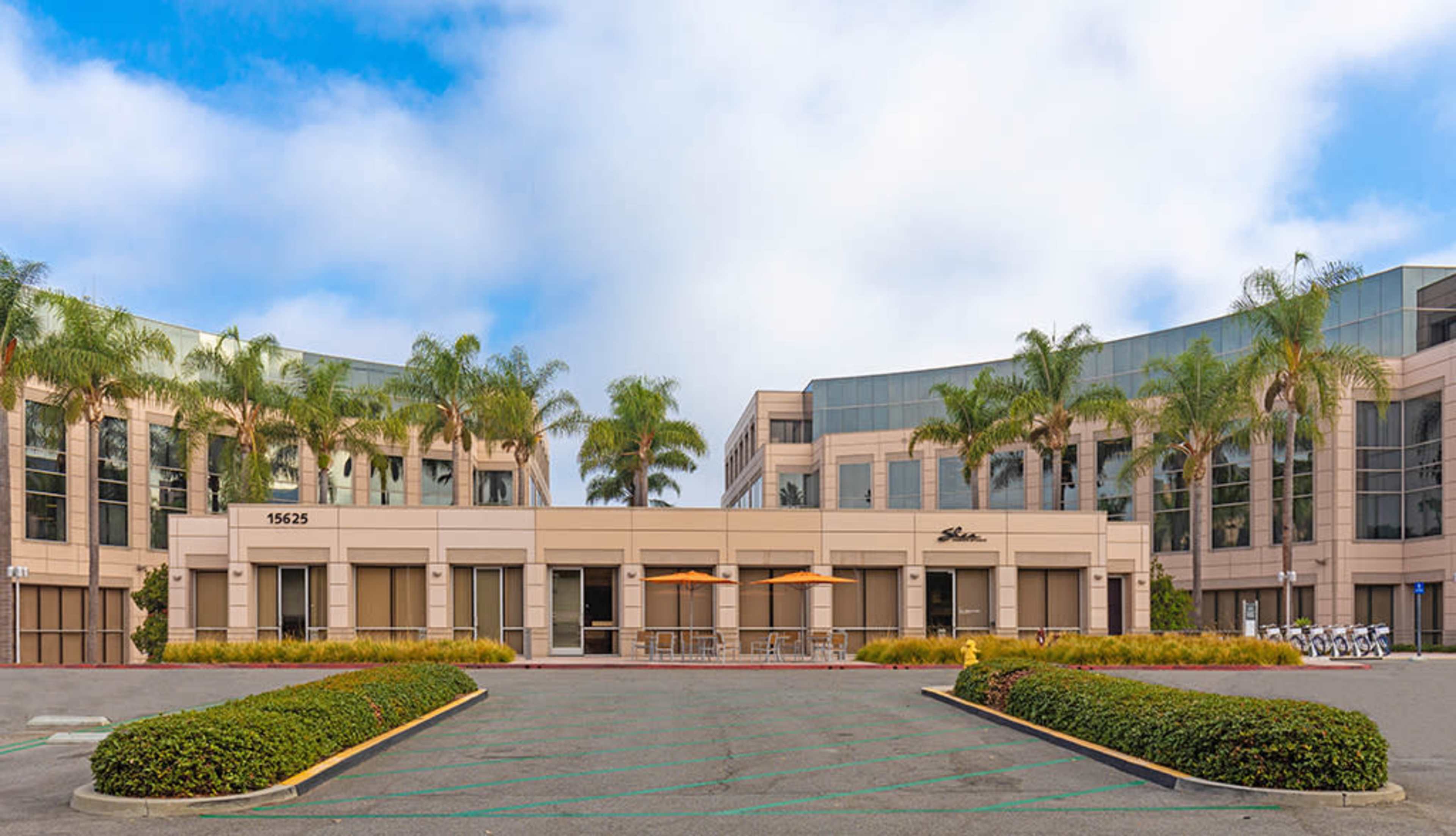 A modern office building complex with palm trees, a parking lot, and outdoor seating under umbrellas.