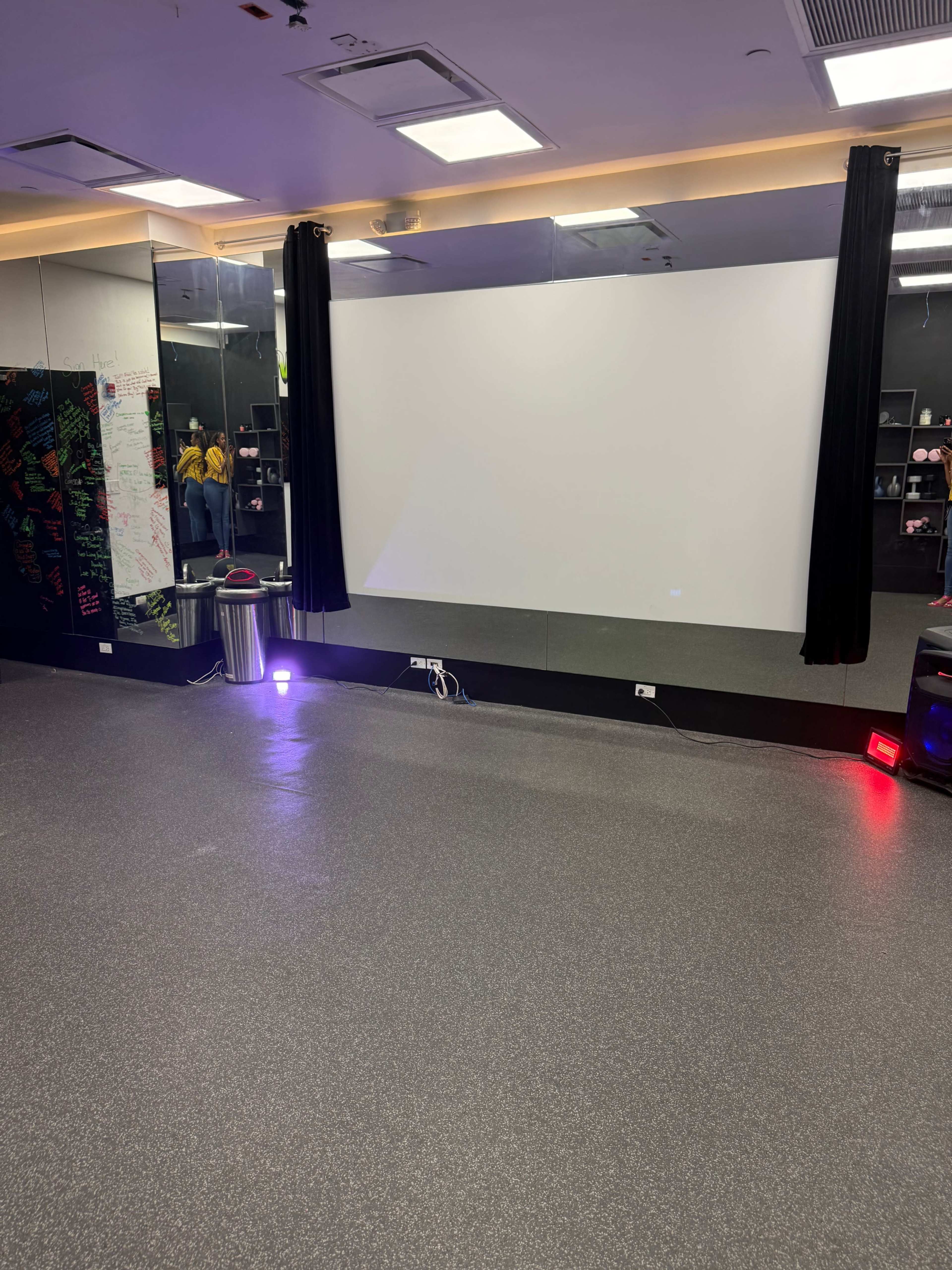 The image shows a fitness studio with a large projection screen, black curtains, and rubber flooring, reflected in a wall of mirrors.