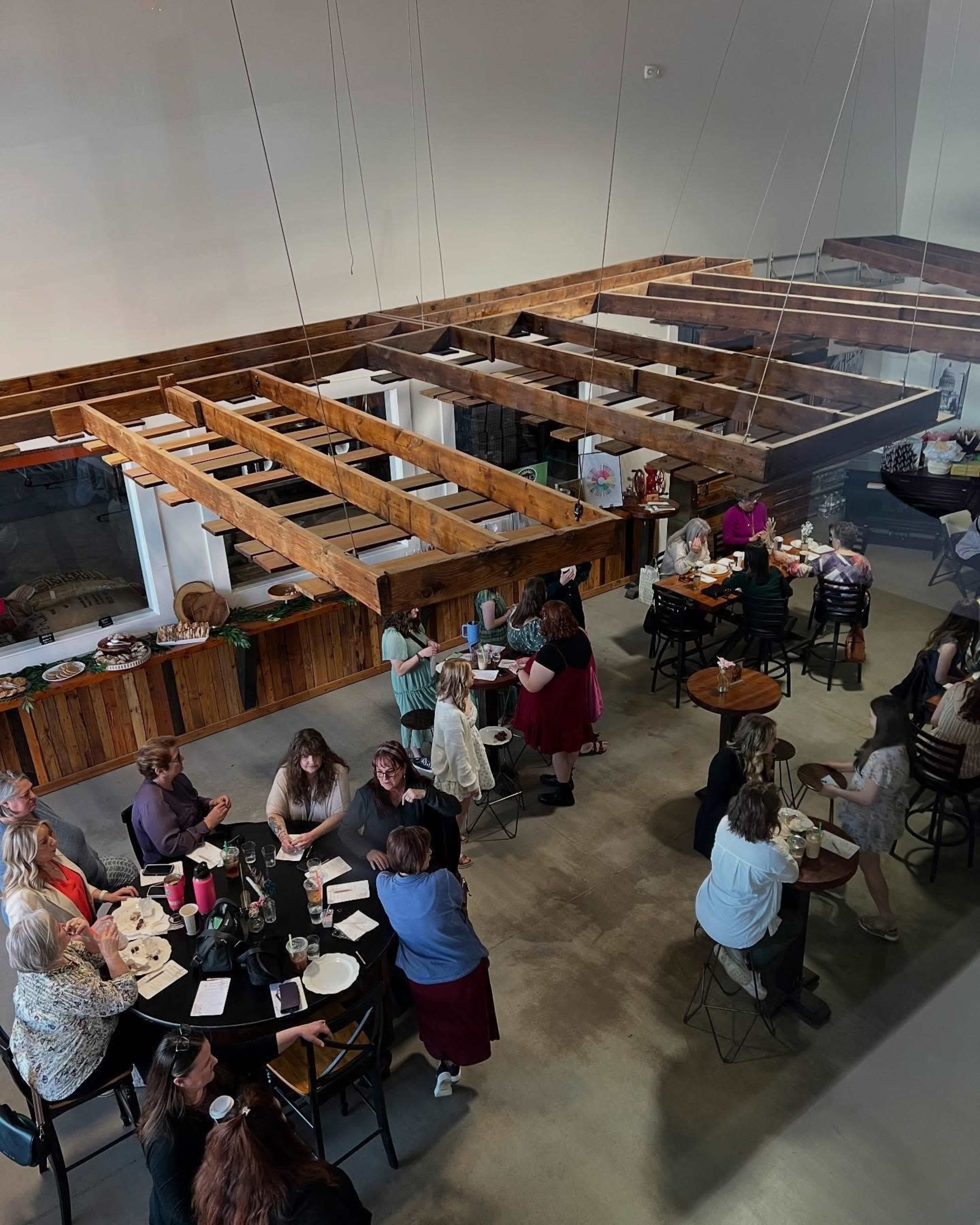 A group of people is gathered in a spacious venue with wooden beams, seated at tables and mingling.
