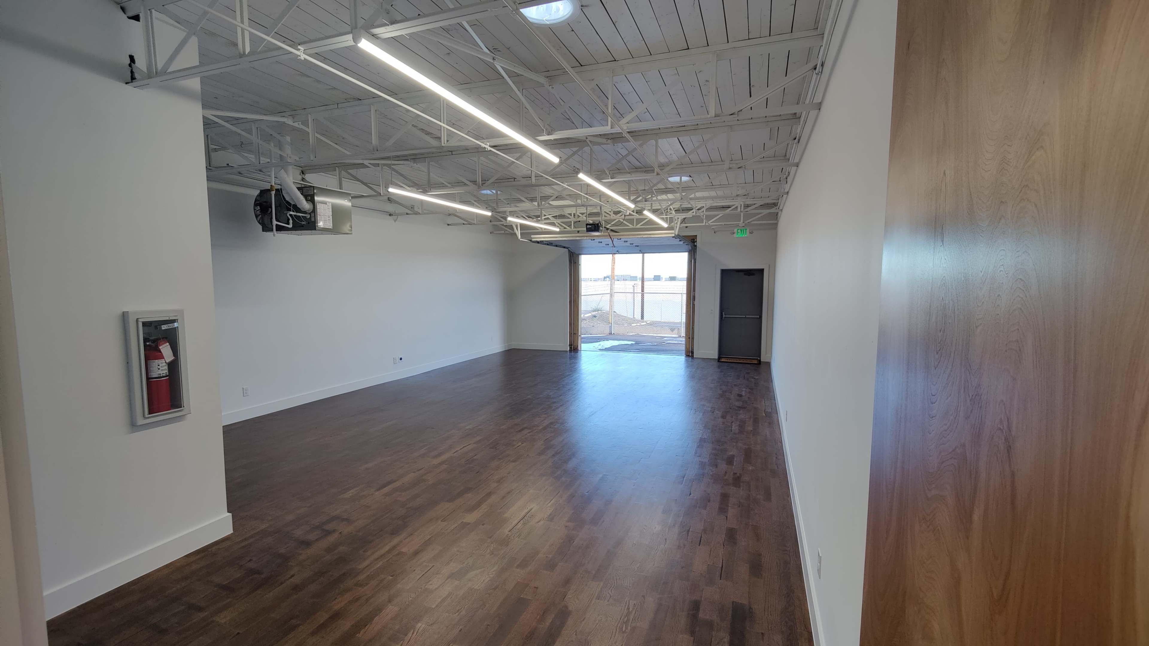 The image shows a spacious, empty room with wooden flooring, white walls, and a large window at one end.