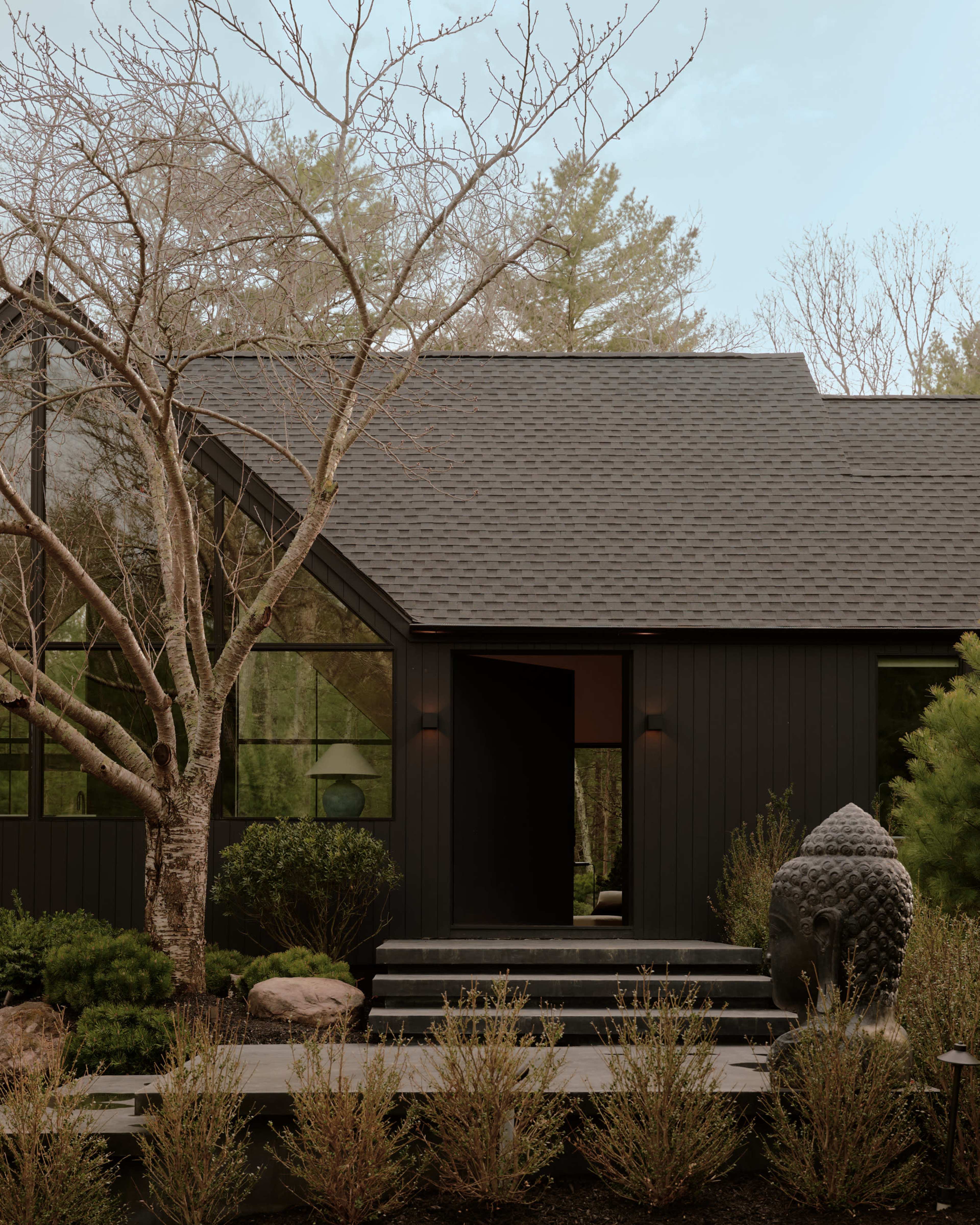 A black-framed modern house with large windows is set among trees and shrubs, featuring a stone pathway and a Buddha statue in the foreground.