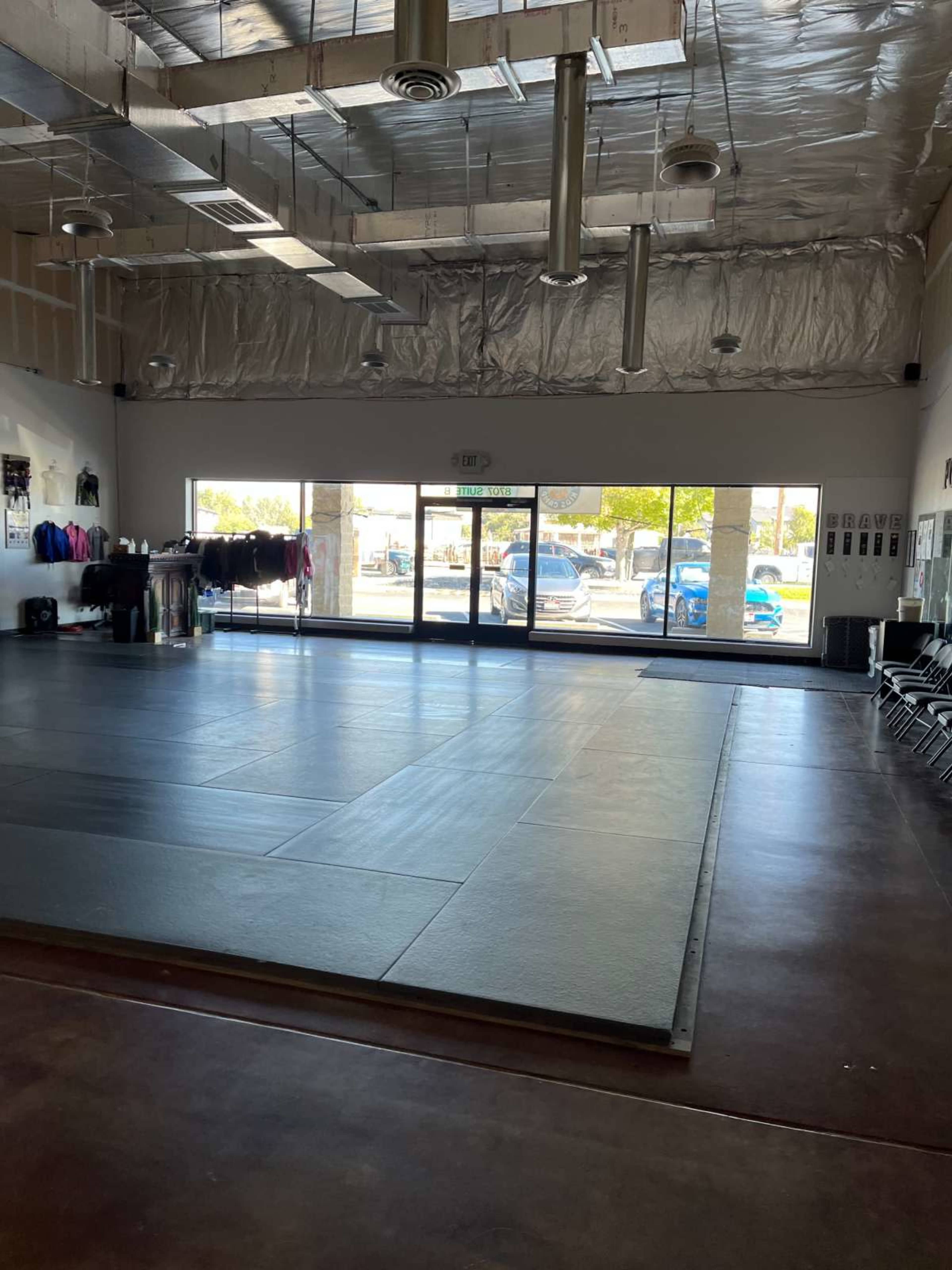 The image shows an empty martial arts studio with a large mat area and large windows letting in natural light.