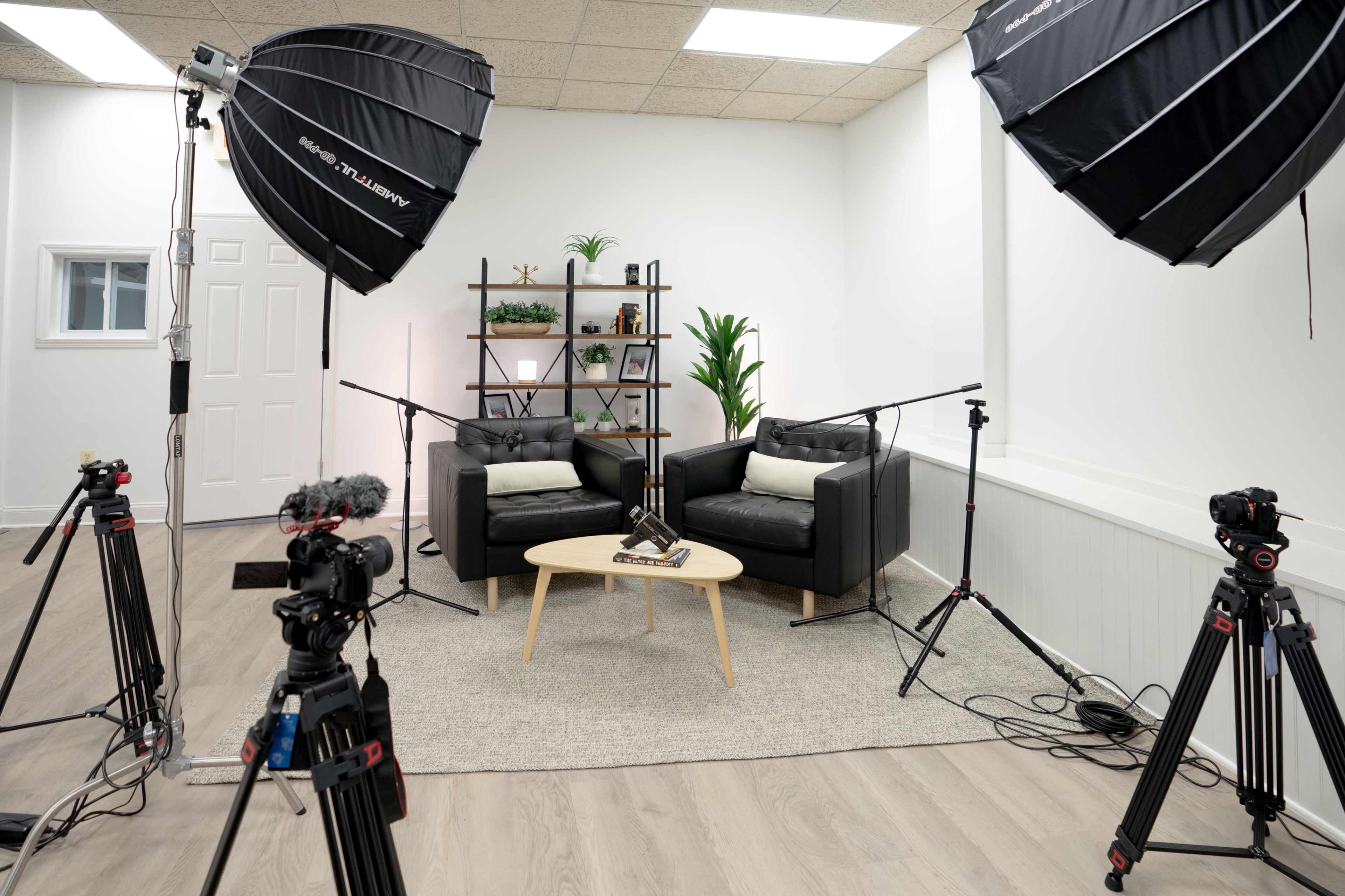 The image shows a modern interview setup with two black armchairs, a small table, and three cameras positioned on tripods, surrounded by large softbox lights in a well-lit studio room.
