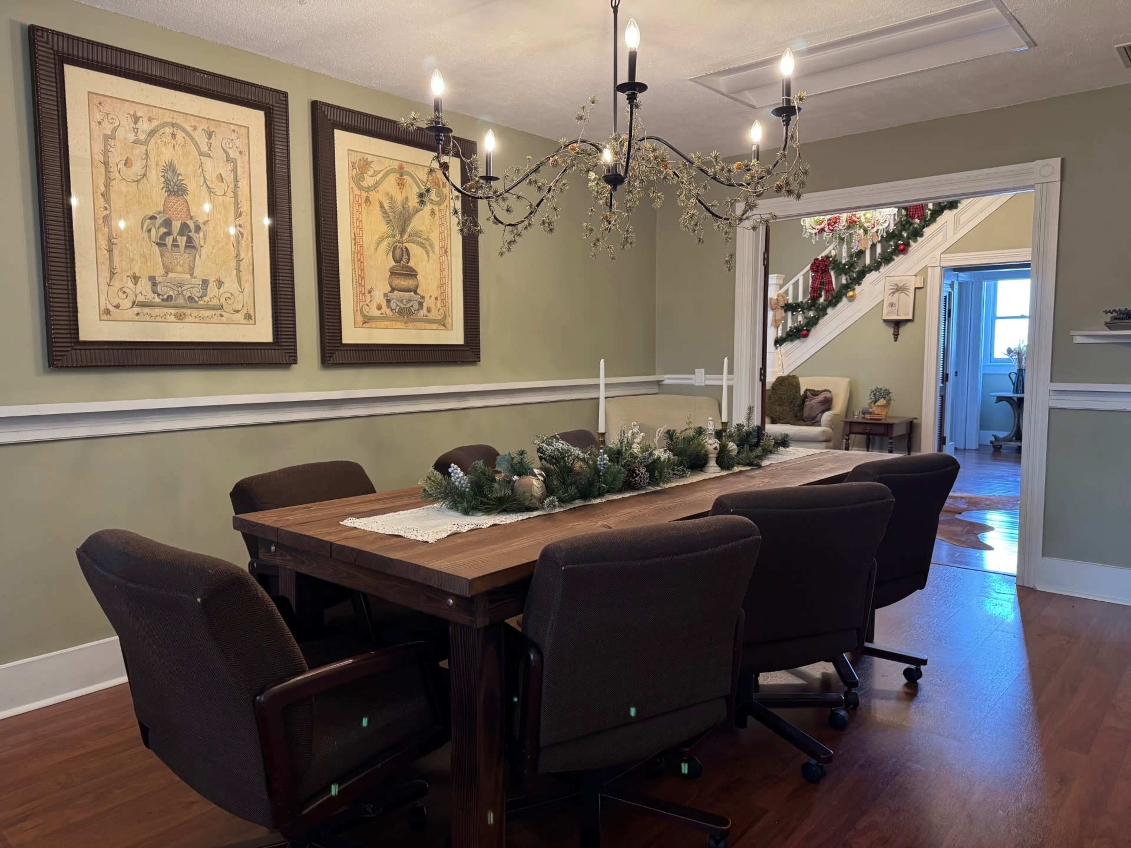 The image shows a dining room with a large wooden table surrounded by chairs, featuring a decorative centerpiece and framed artwork on the walls.