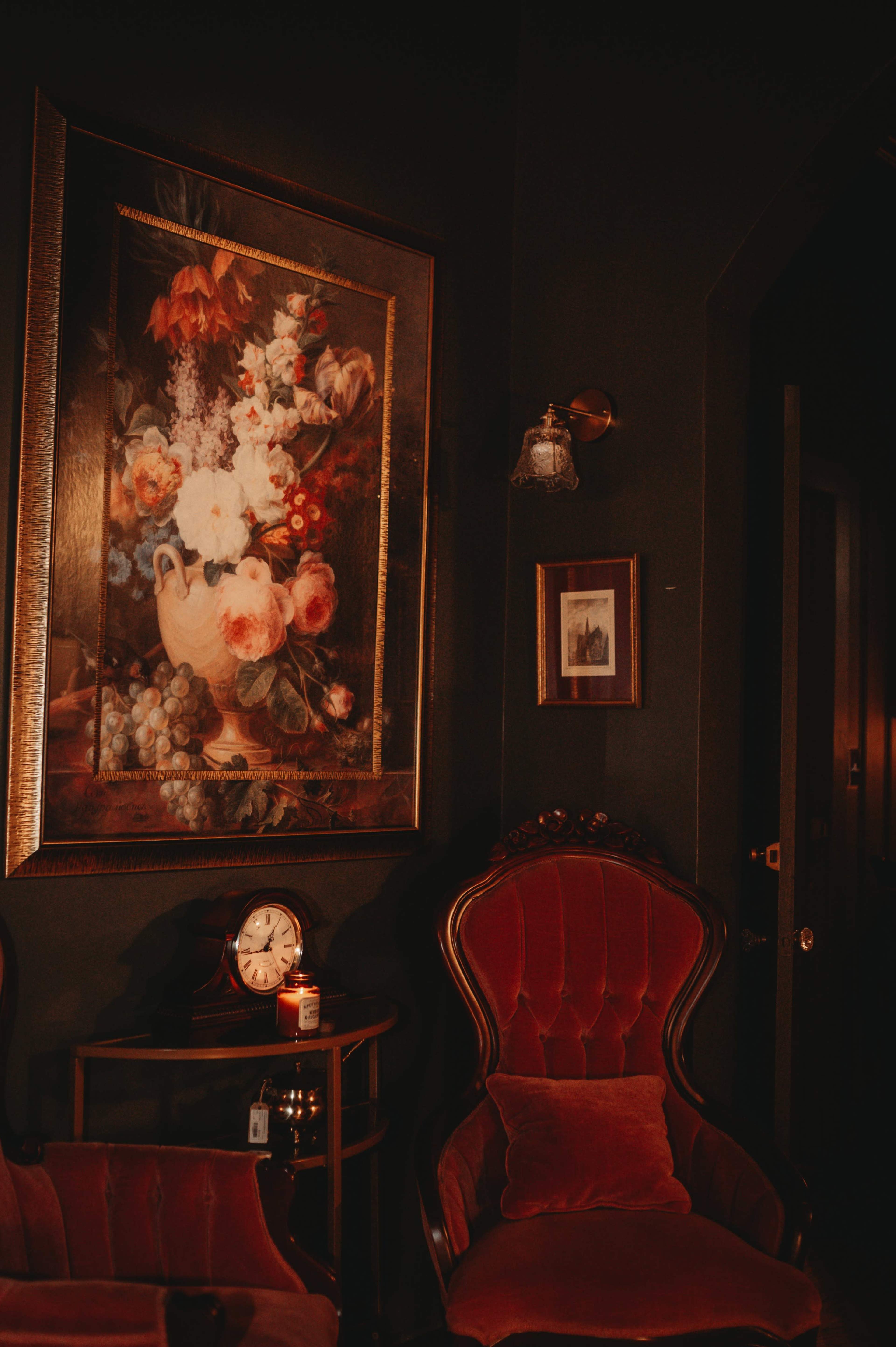 The image shows a cozy room with a large floral painting on the dark wall, an antique armchair, a vintage clock, and a candle on a side table.