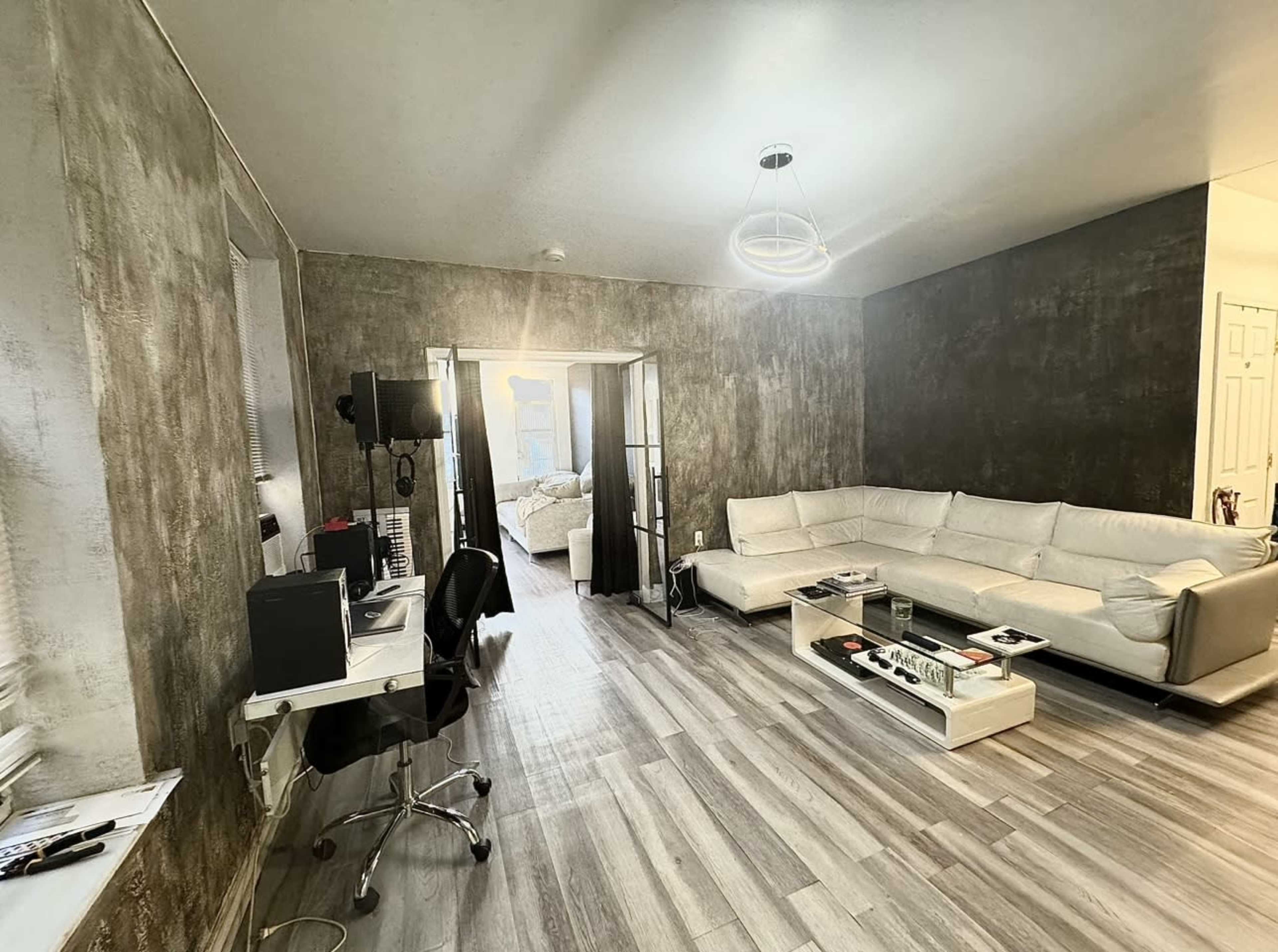 A living room with gray walls, a white sectional sofa, a desk with a computer, and light-colored wooden flooring leading to a doorway.