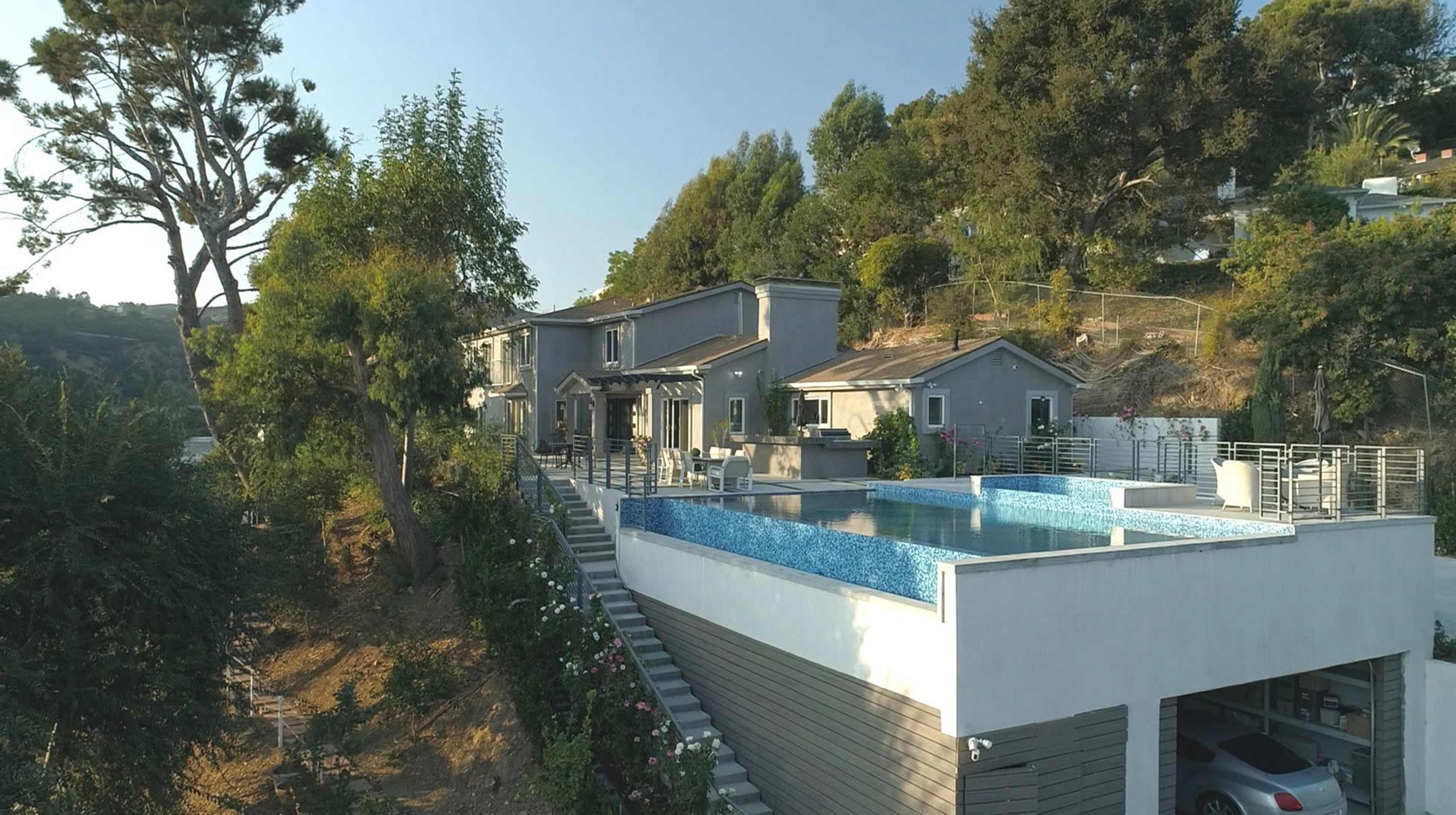 A modern two-story house with a large swimming pool and landscaped garden overlooks a hillside.