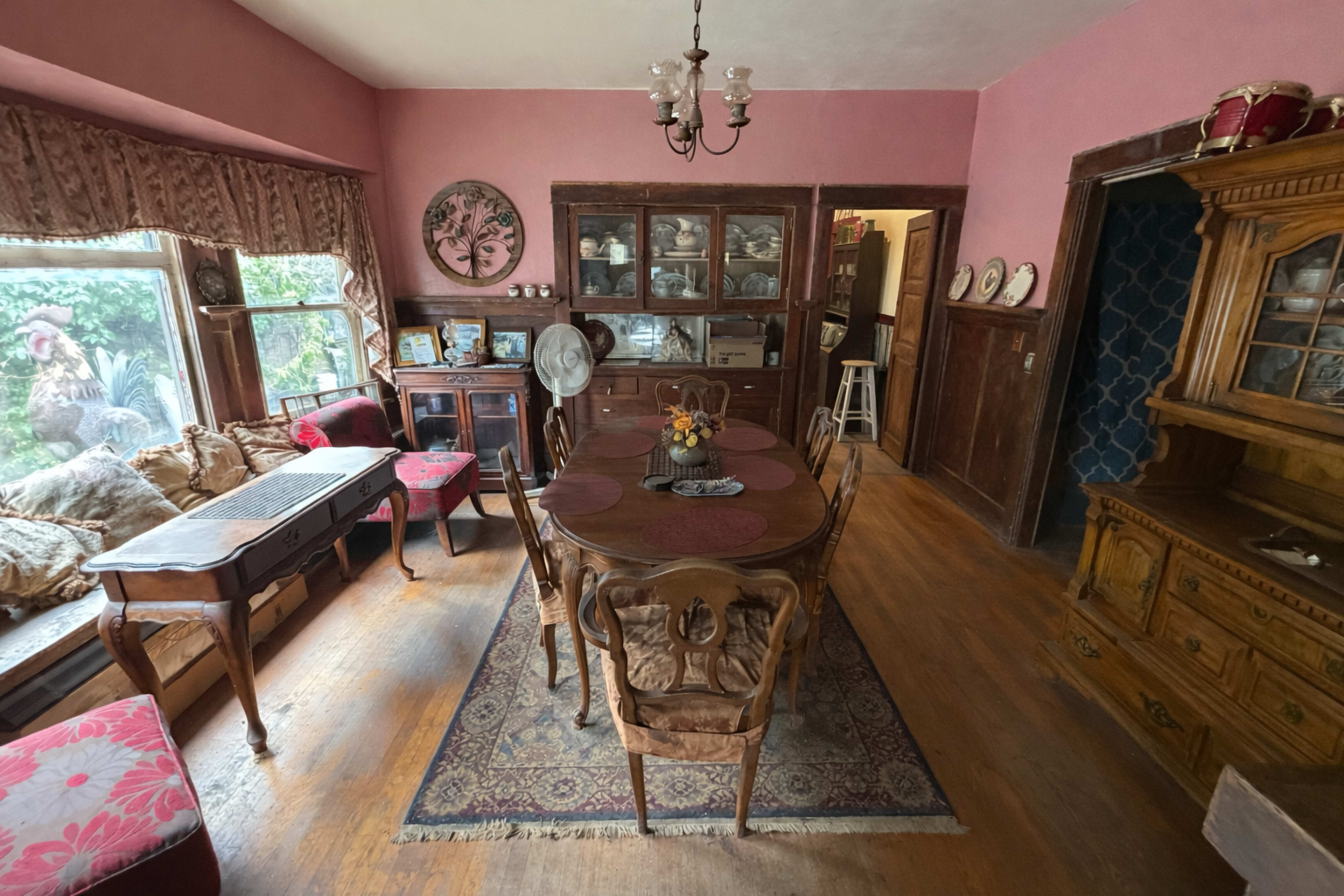 The image shows a vintage dining room with a wooden table, several chairs, a cabinet displaying dishware, and large windows overlooking a garden.