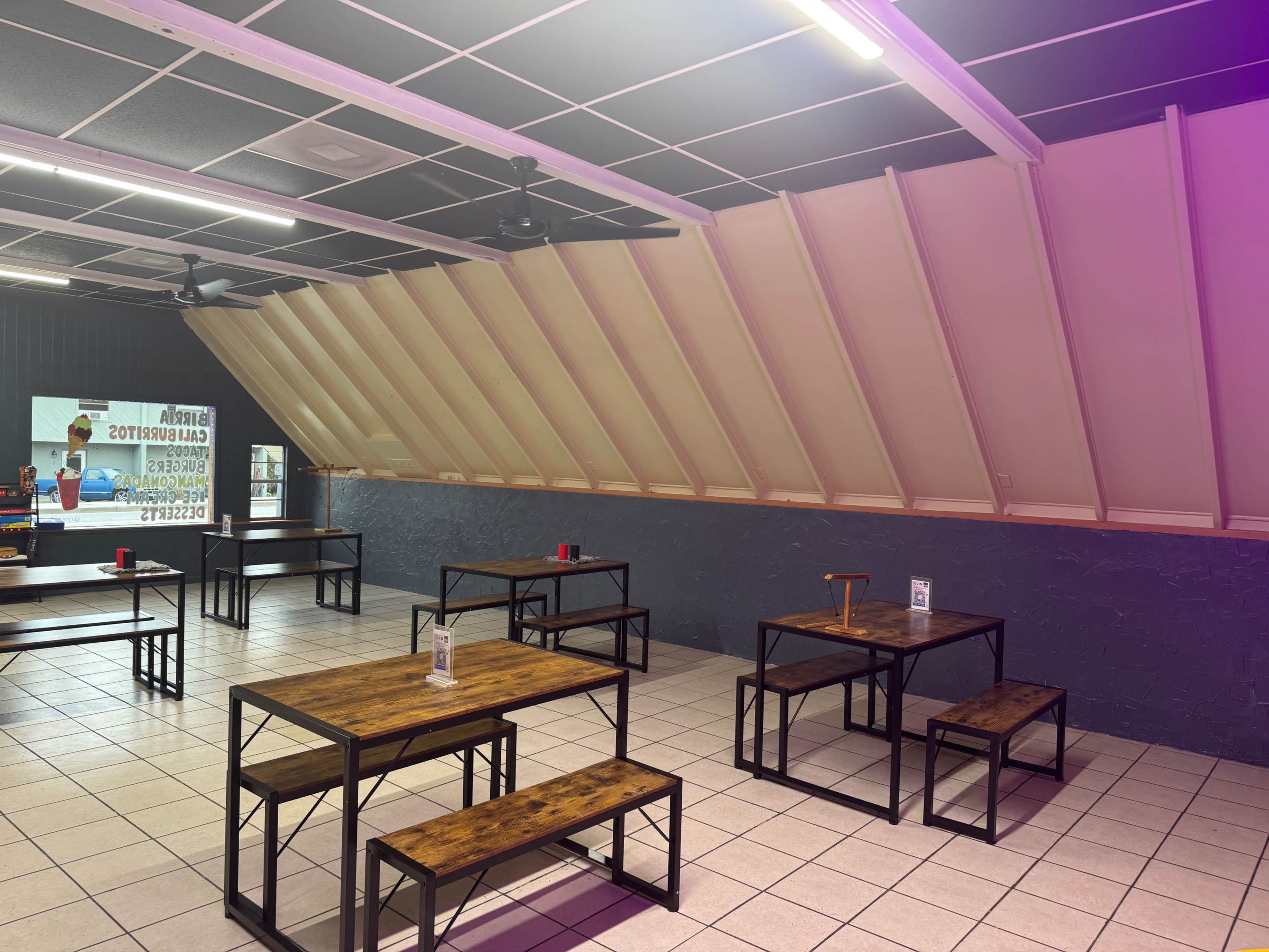 The image shows an indoor dining area with several metal-framed tables and benches arranged on a tiled floor, under a sloped roof with a screen displaying advertisements on one wall.