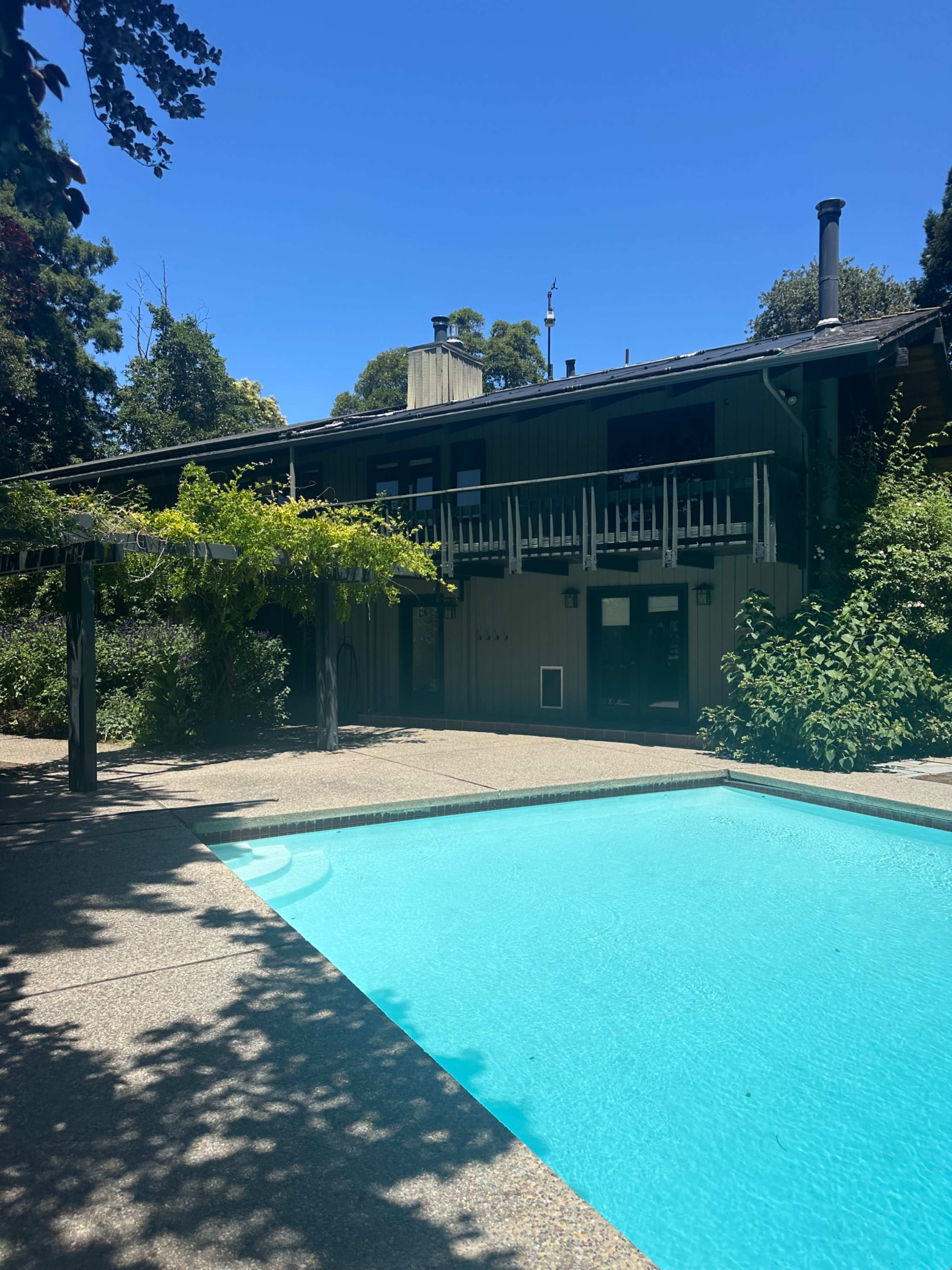 The image shows a swimming pool surrounded by a patio and greenery, with a two-story house visible in the background against a clear blue sky.