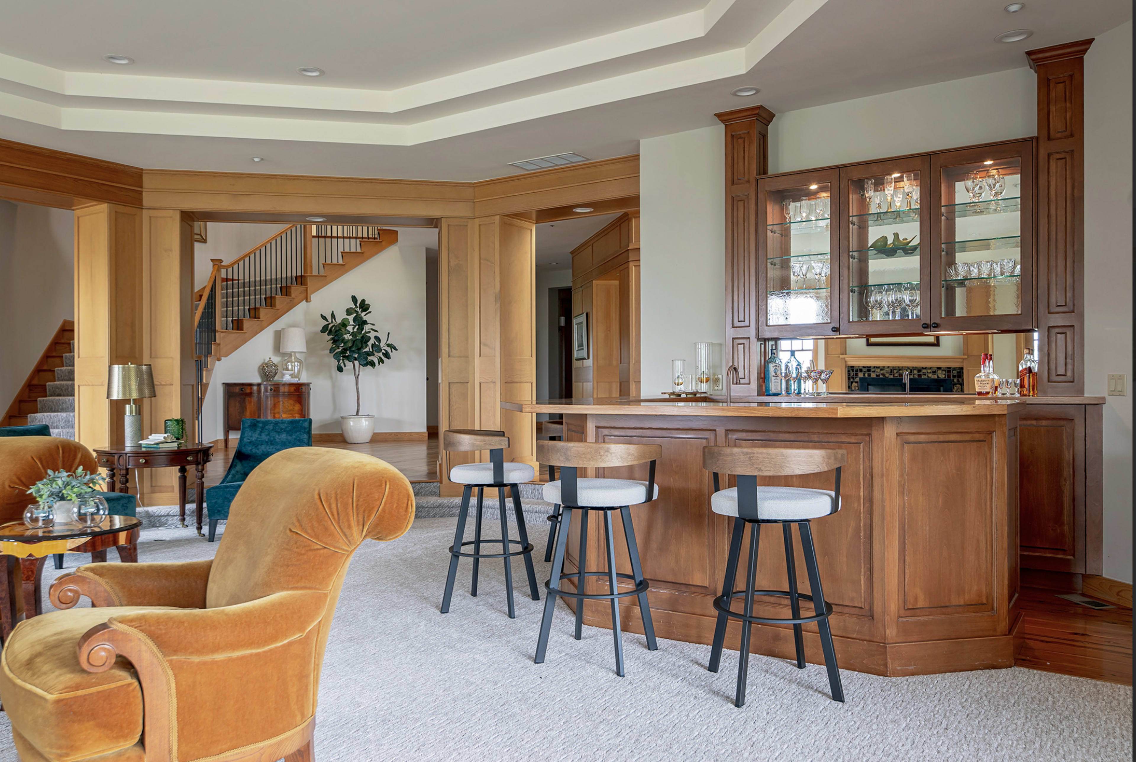The image shows a spacious living area with a bar, featuring wooden cabinetry, several stools, and a staircase in the background.