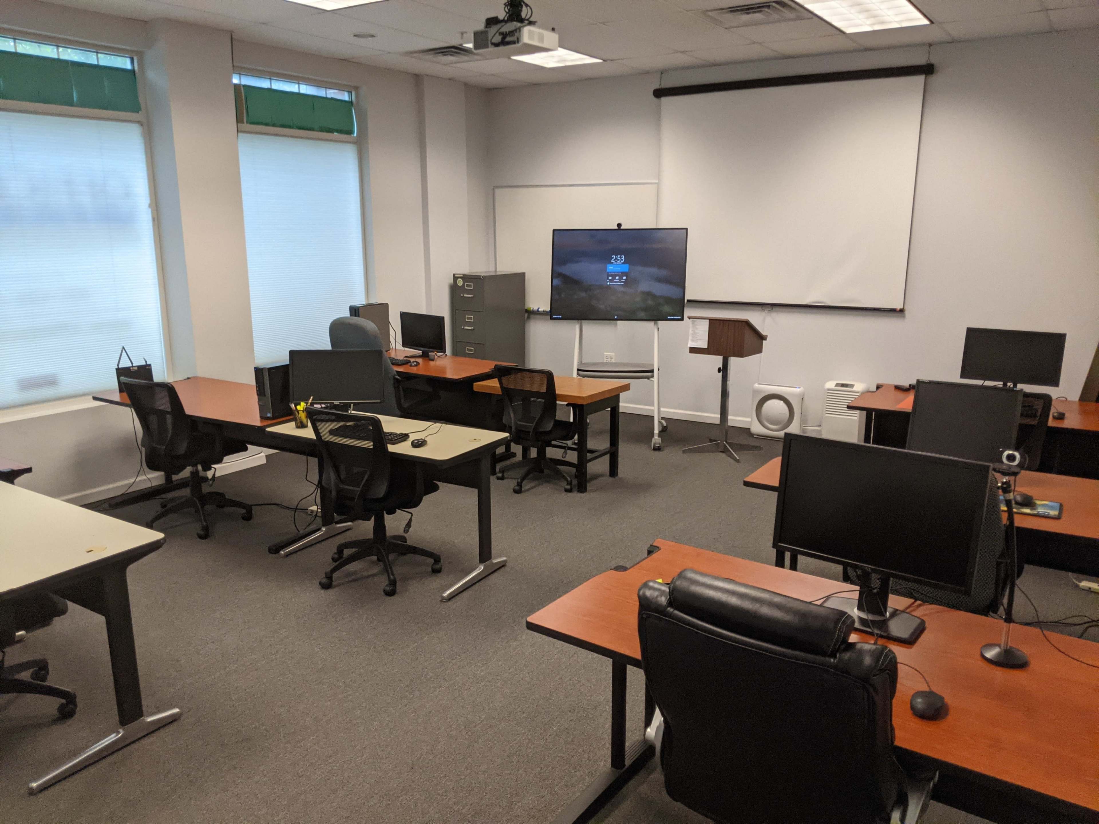 A spacious classroom or conference room with several desks arranged in rows, each equipped with computers, and a large screen mounted on a stand at the front.