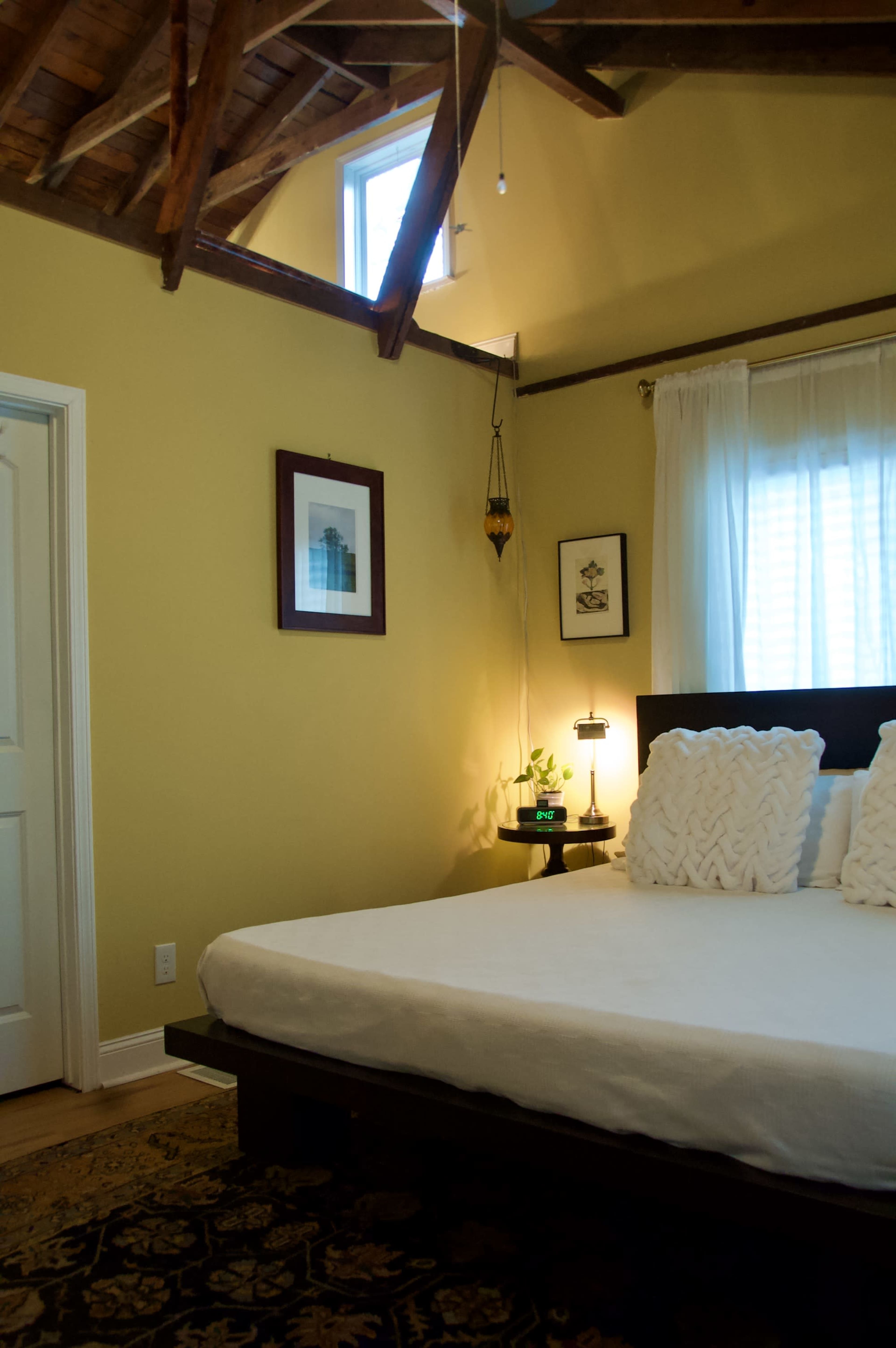 The image shows a cozy bedroom with a wooden bed, two decorative pillows, and a window allowing natural light to enter, along with a small nightstand and framed artwork on the wall.