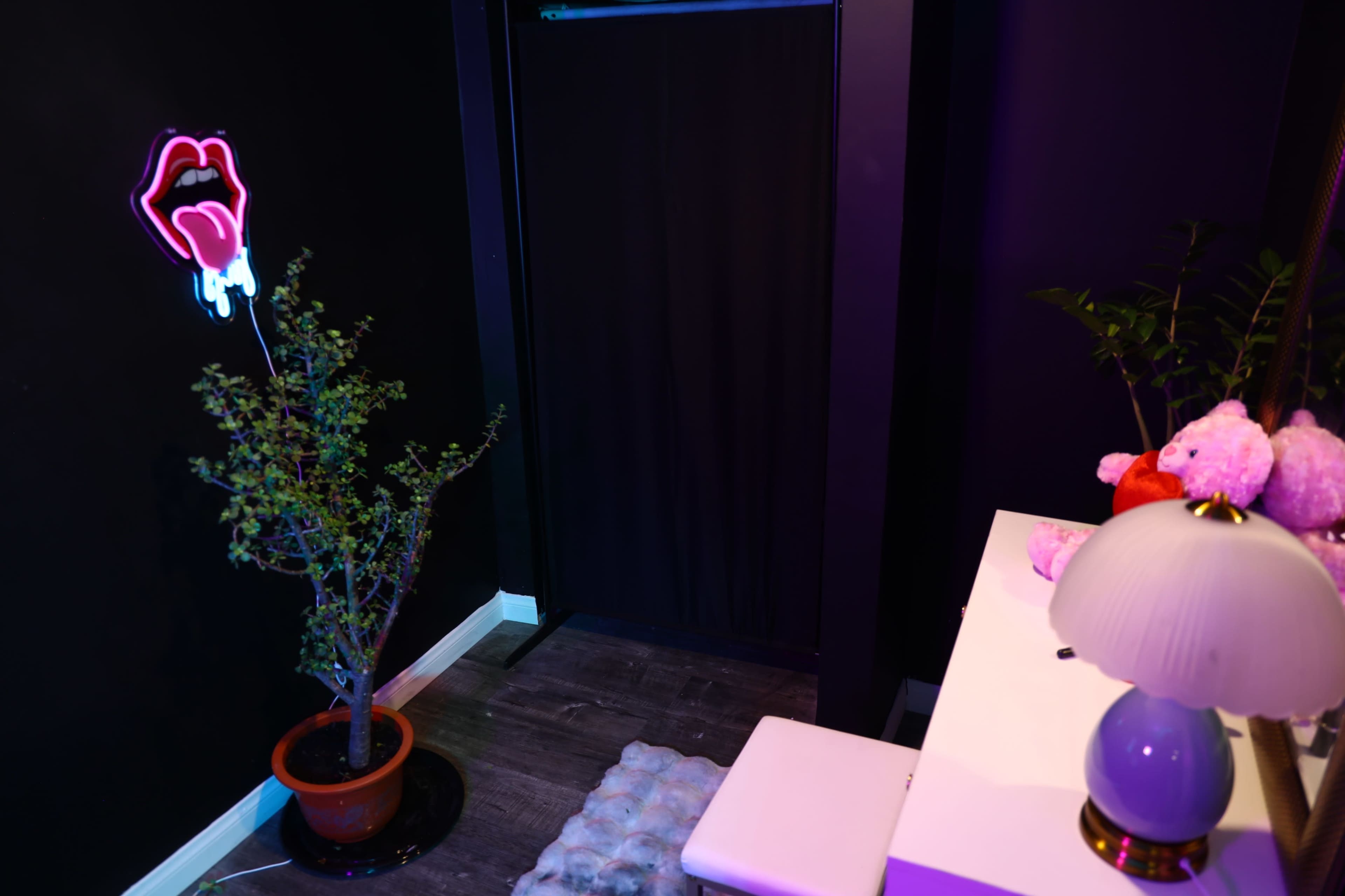 The image depicts a small, dimly lit room with a black wall, a neon lips sign, a potted plant, and a white table with a lamp.