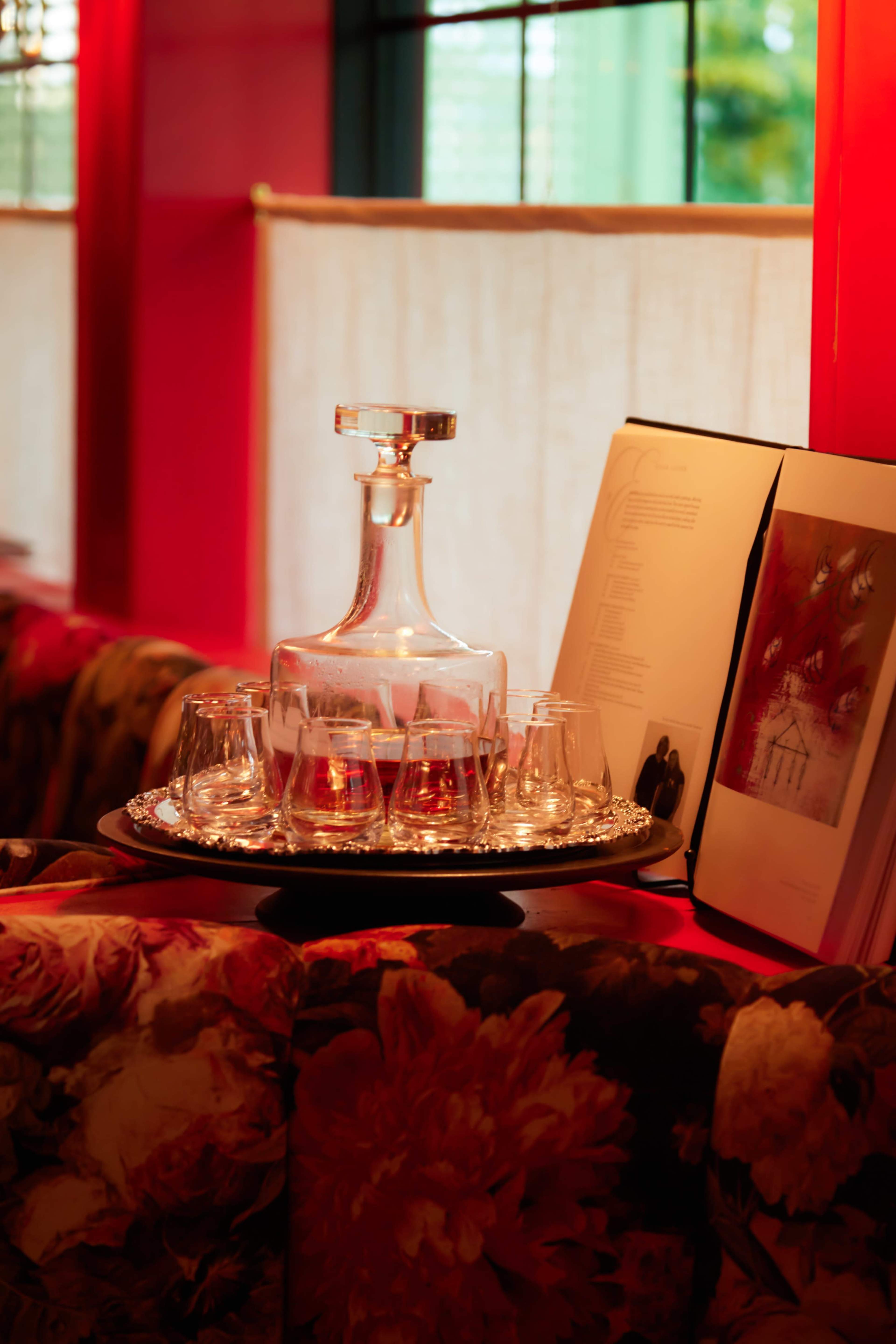 A glass decanter is placed on a silver tray surrounded by glasses and art on a floral-print sofa against a vibrant pink wall.