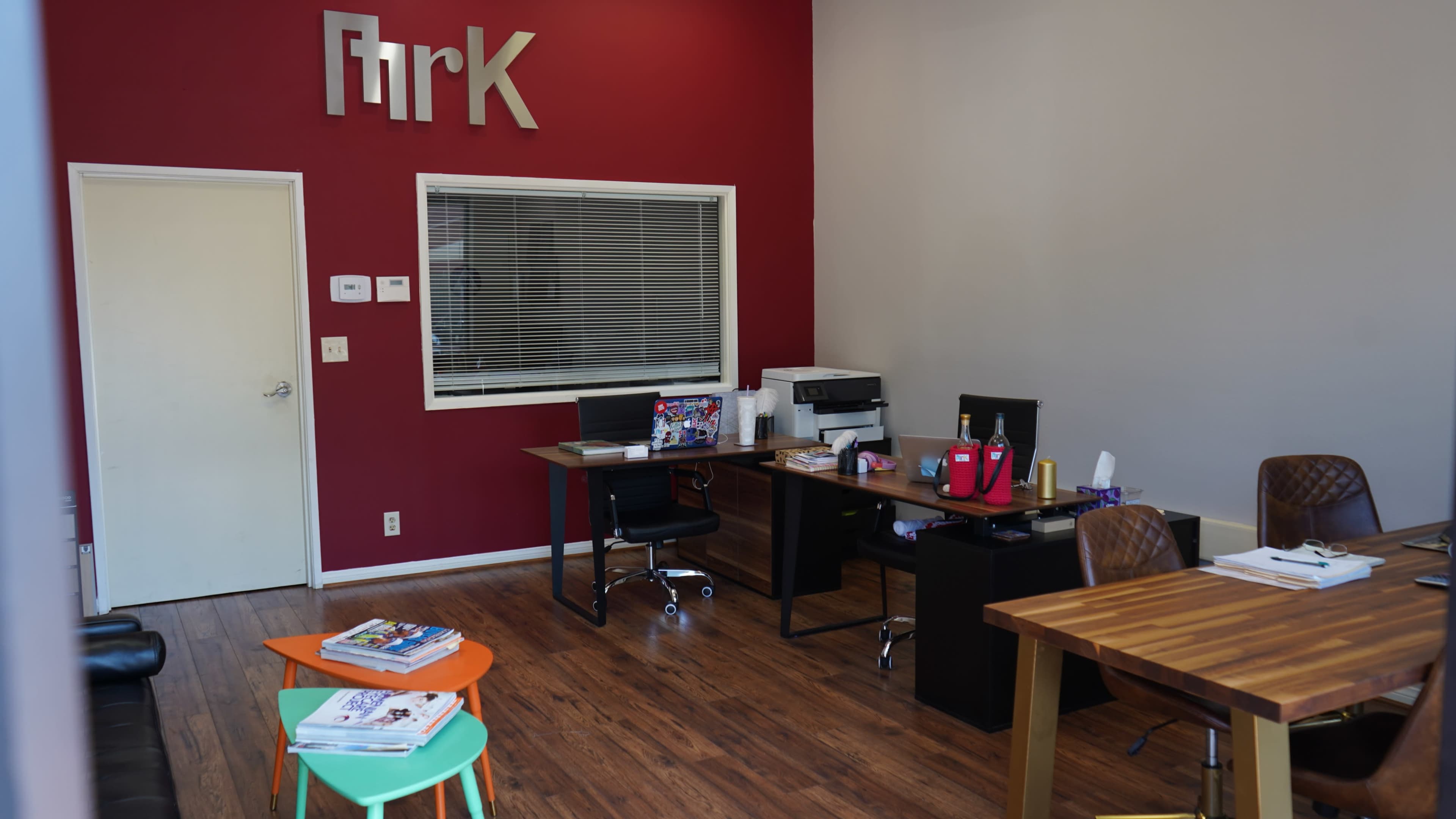 The image shows an office space with two desks, a printer, and a colorful coffee table in front of a red accent wall.