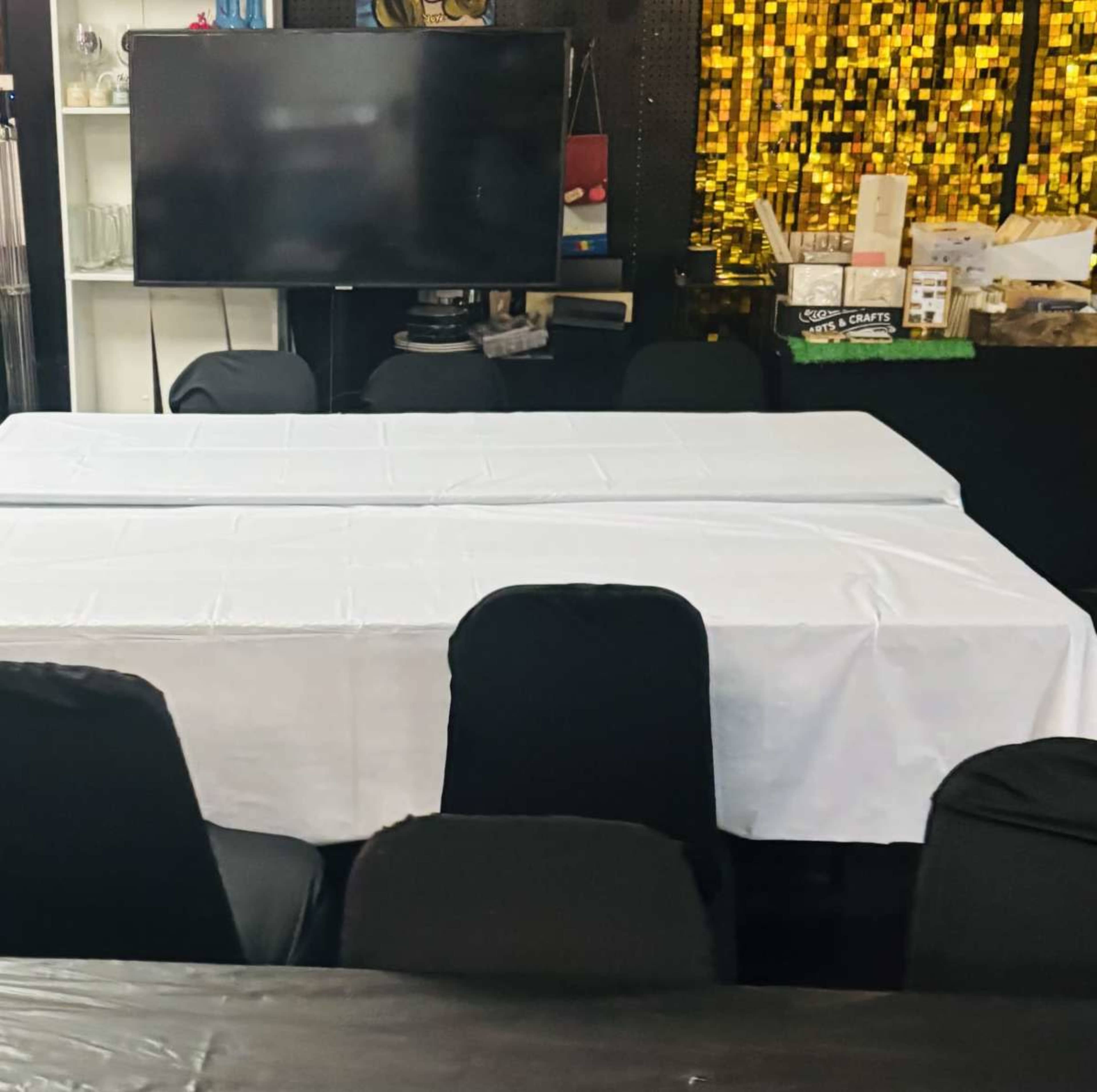 The image shows a meeting or event space with two white-covered tables, black chairs, and a large television screen in the background.