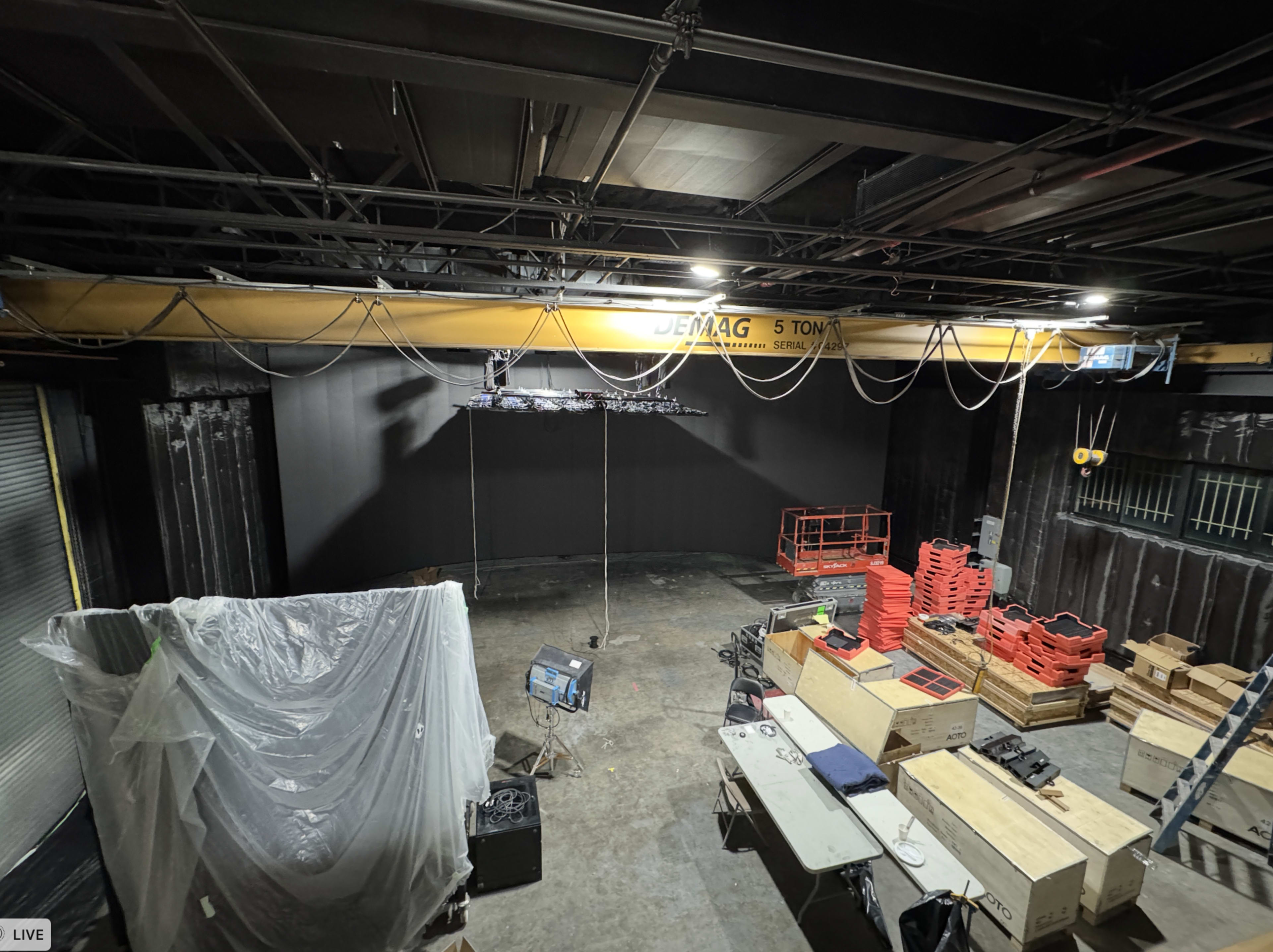 The image shows an industrial space with a high ceiling, featuring a large overhead crane, various tools and equipment scattered on the floor, and a blank wall in the background.