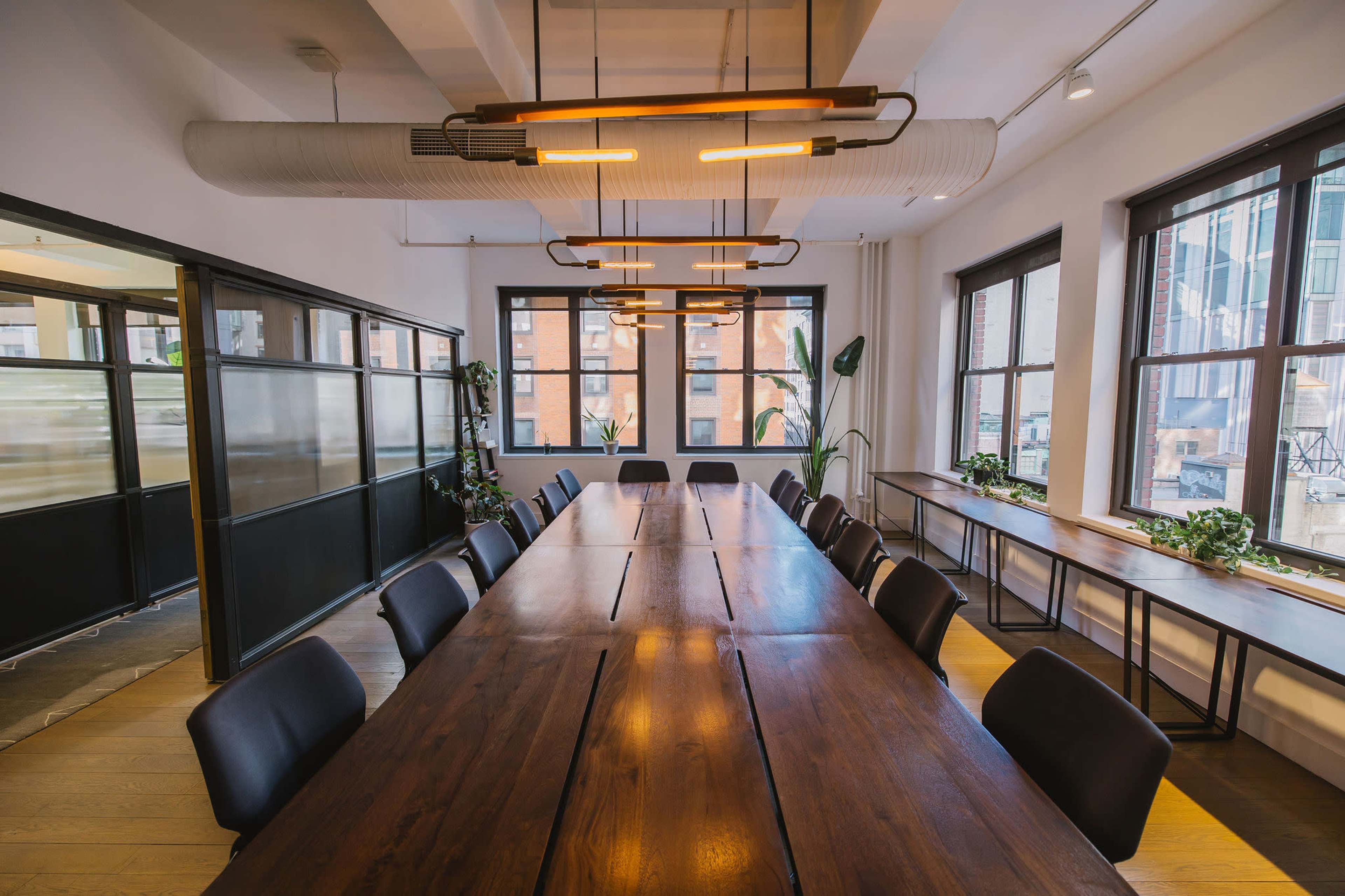 A large wooden table with black chairs occupies a well-lit conference room featuring multiple windows and modern lighting.