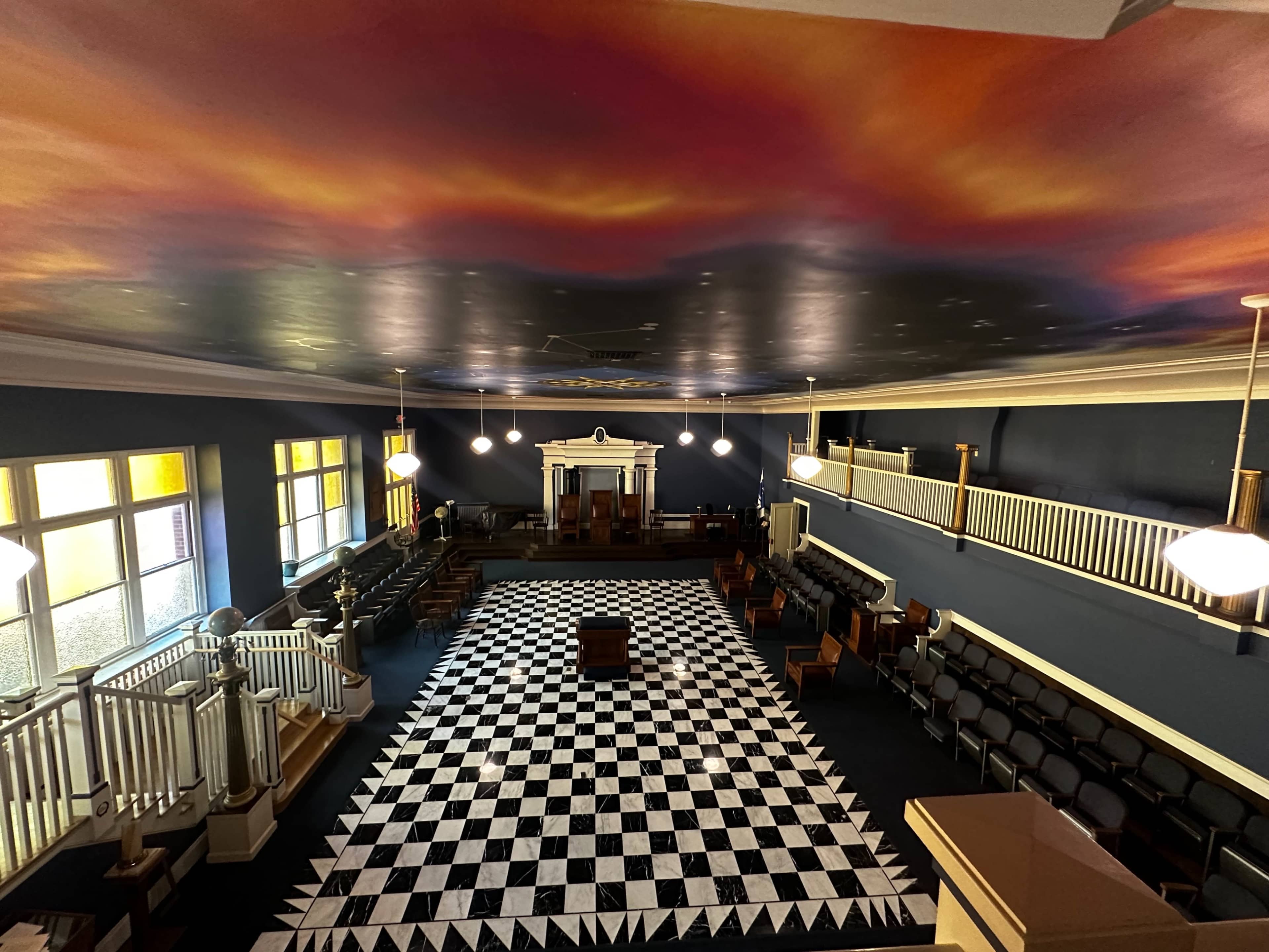 The interior of a large hall features a black and white checkered floor, rows of seating, and a painted ceiling that resembles a sunset.