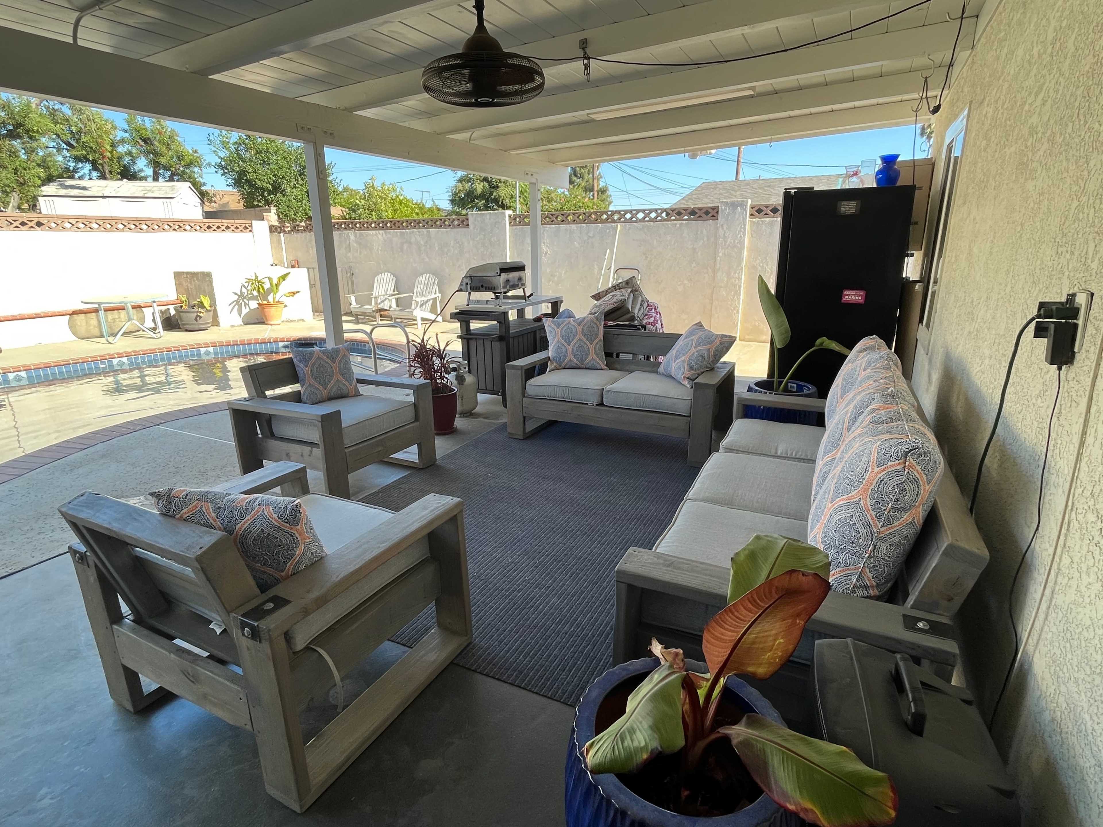 The image shows a covered patio with wooden furniture arranged around a rug near a pool.