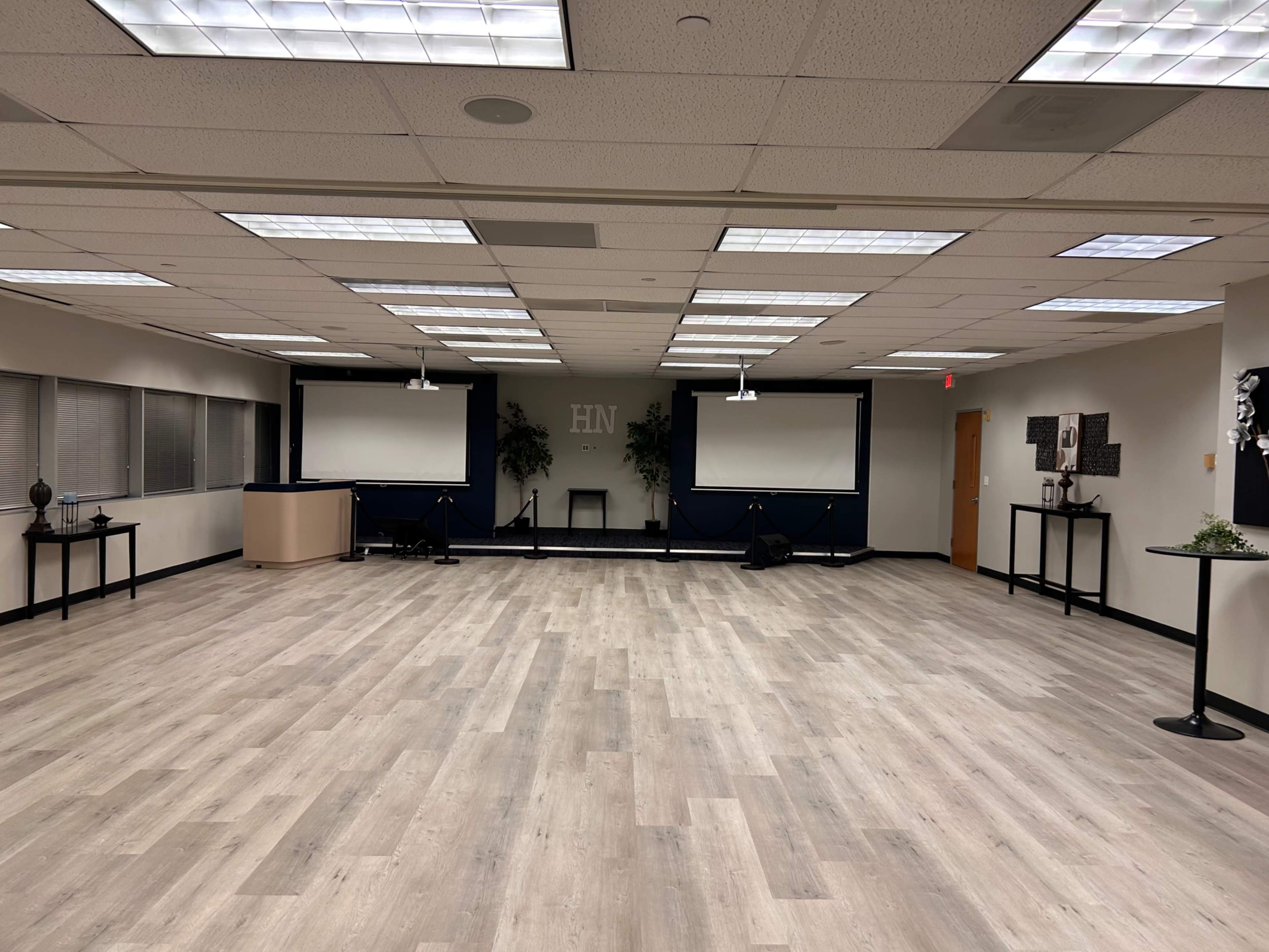 The image shows a spacious, minimally decorated room with two screens at the front and several tables arranged around the perimeter.