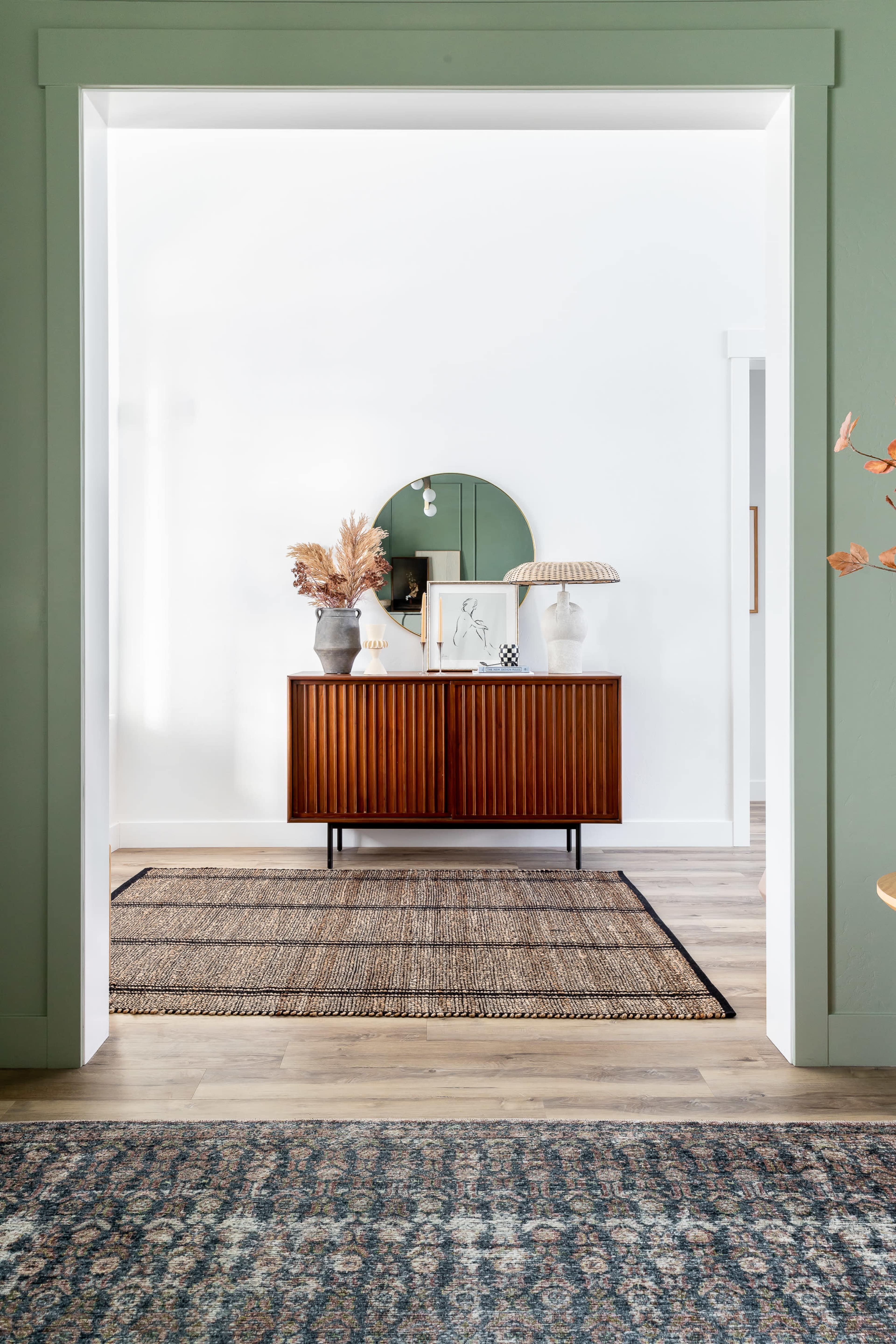 A narrow entryway features a wooden sideboard with decorative items and a round mirror above it, framed by green walls and a woven rug on the floor.